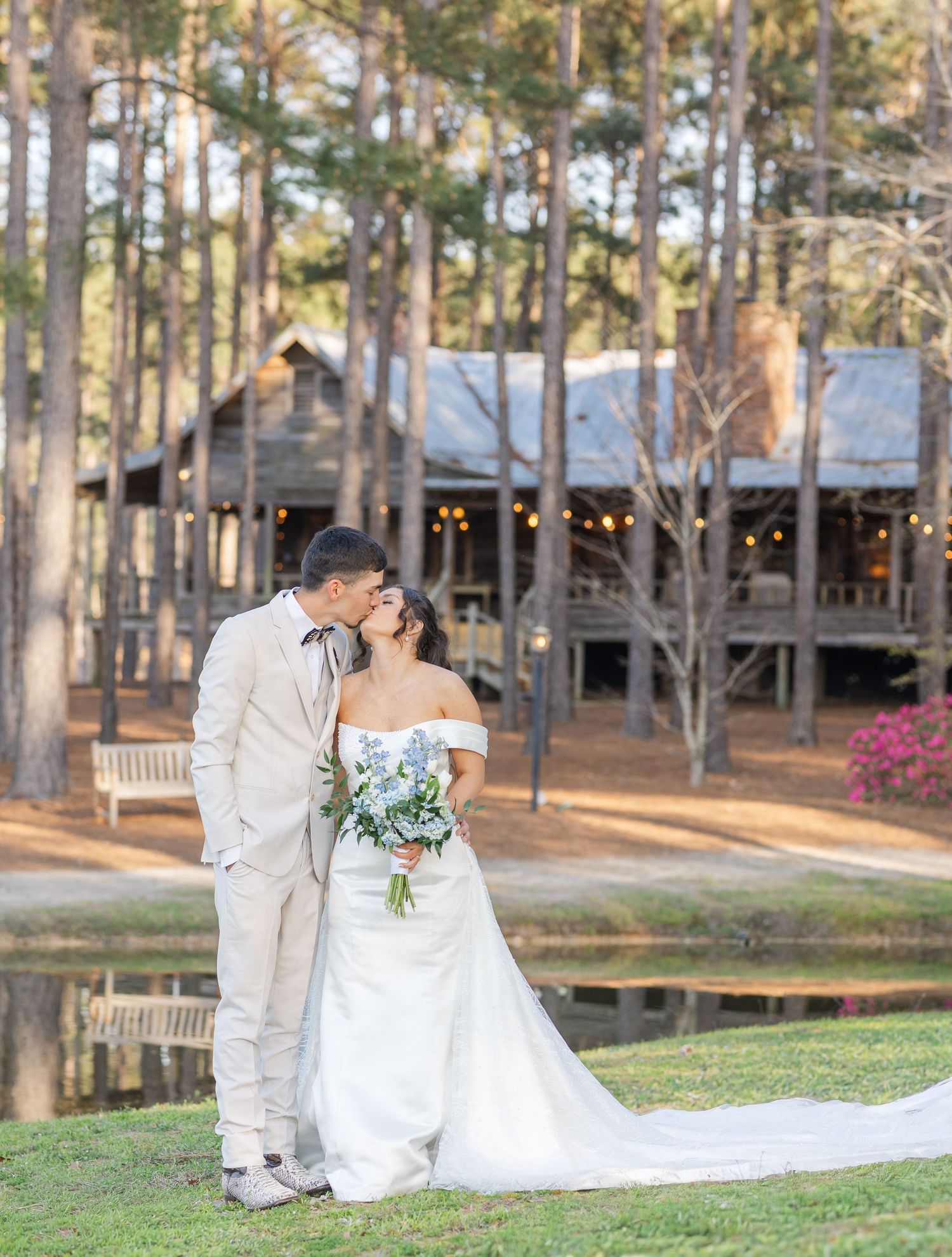 Bride and groom share a kiss near a pond with a rustic log cabin reception venue glowing in golden hour light.