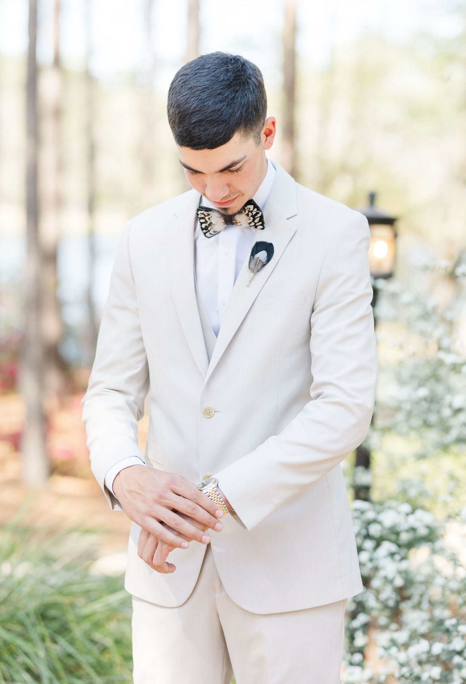 Groom in cream suit adjusting cufflinks with leopard print bow tie and boutonniere at outdoor wedding.