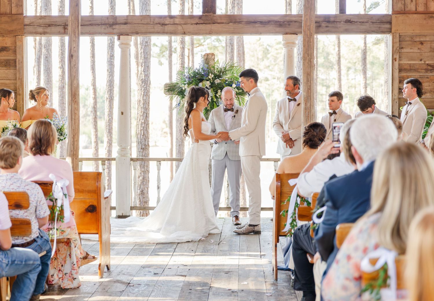 Bride and groom exchange vows at rustic barn wedding ceremony with seated guests watching.