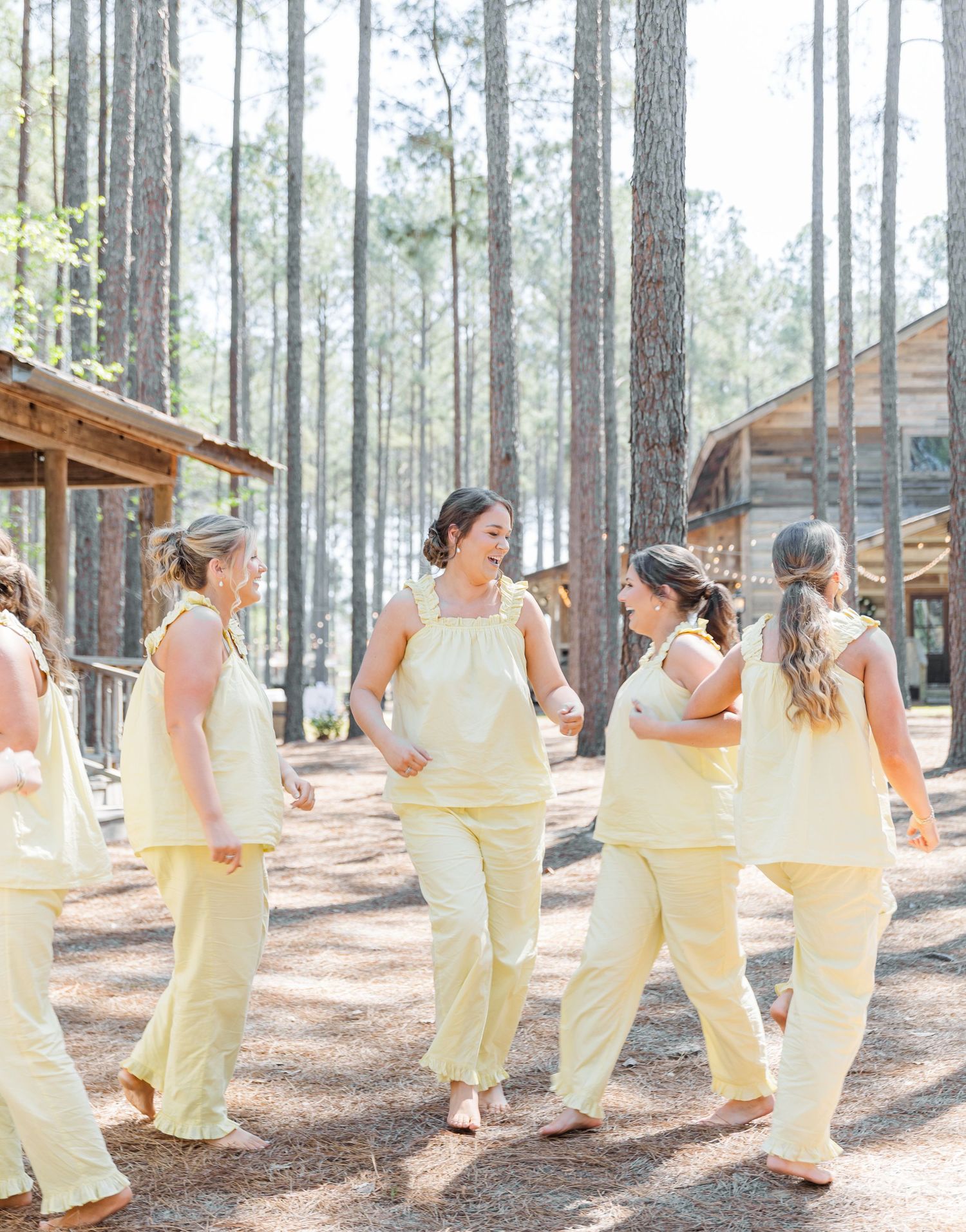 Bridesmaids in yellow outfits walking and laughing together among pine trees at an outdoor wedding venue.