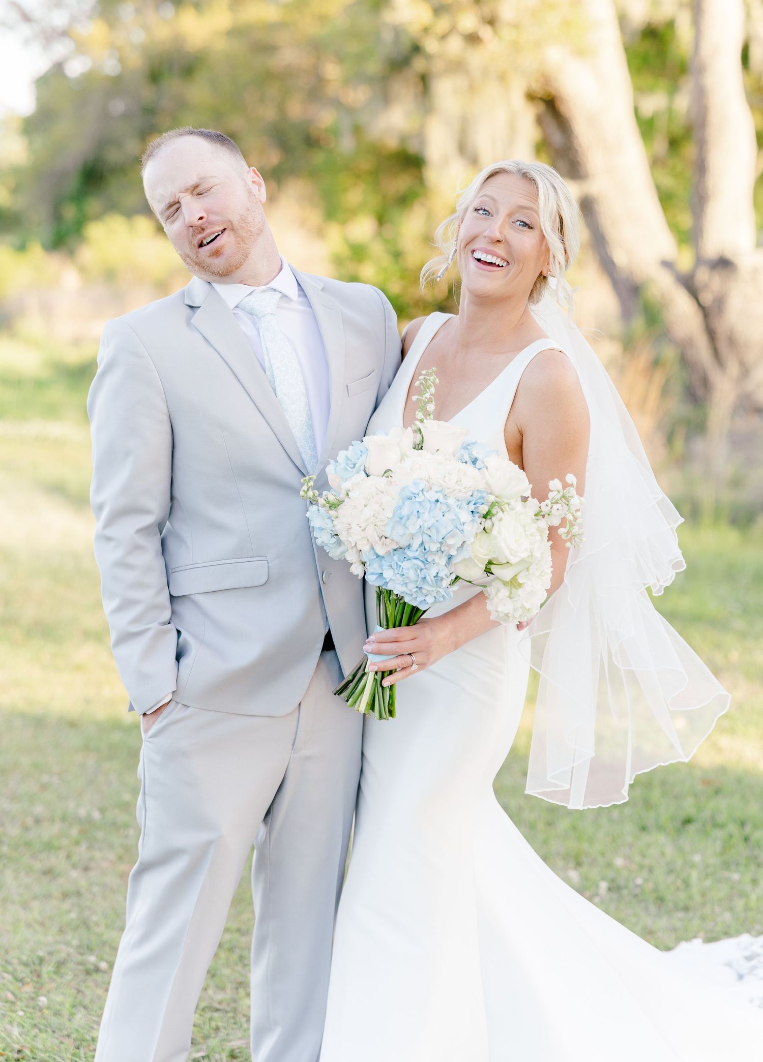 Laughing bride in fitted white gown holding blue hydrangea bouquet standing with groom in light gray suit outdoors.