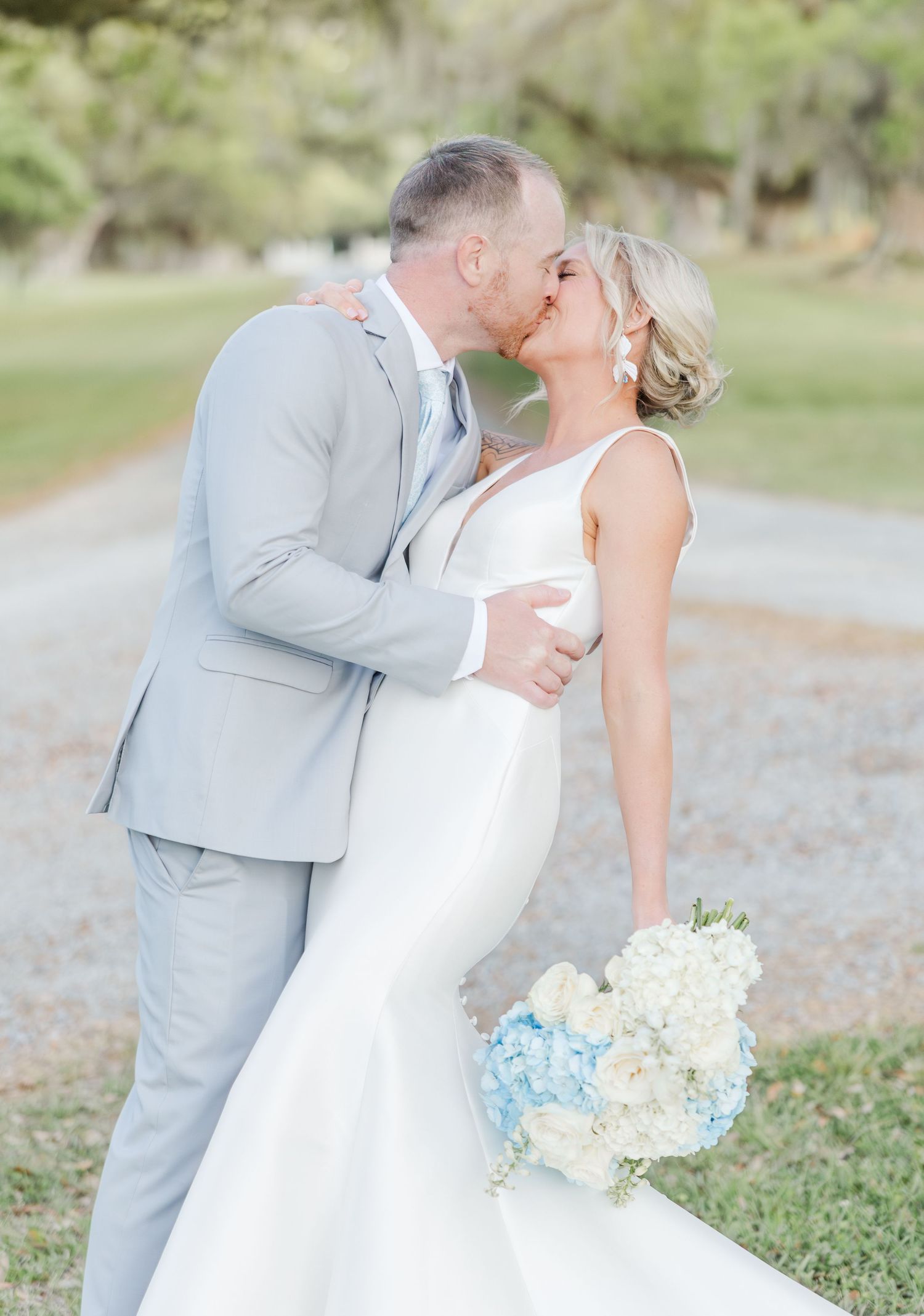 Bride in mermaid gown and groom in gray suit sharing a kiss, holding blue hydrangea bouquet outdoors.