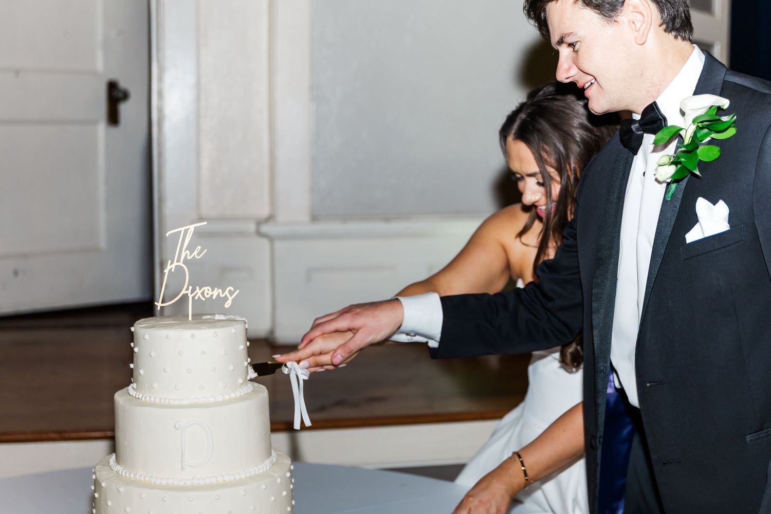 Bride and groom cutting their three-tier white wedding cake topped with The Dixons cake topper at their reception.