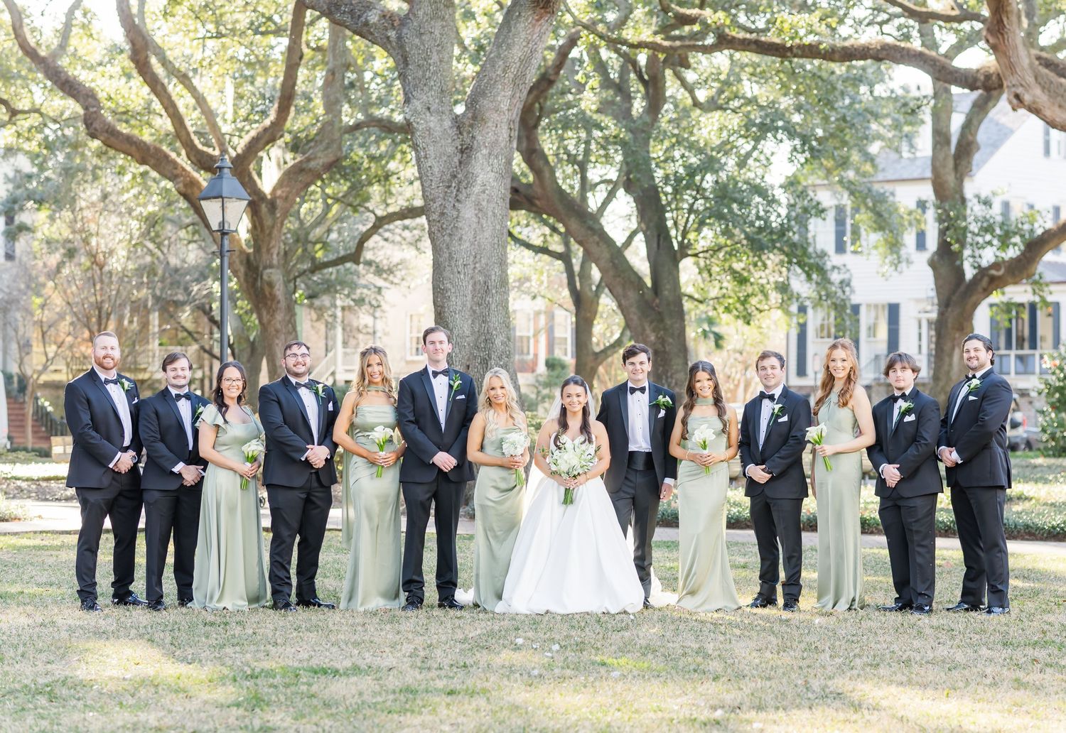 Large wedding party in sage bridesmaid dresses and black tuxedos poses under oak trees at outdoor ceremony.