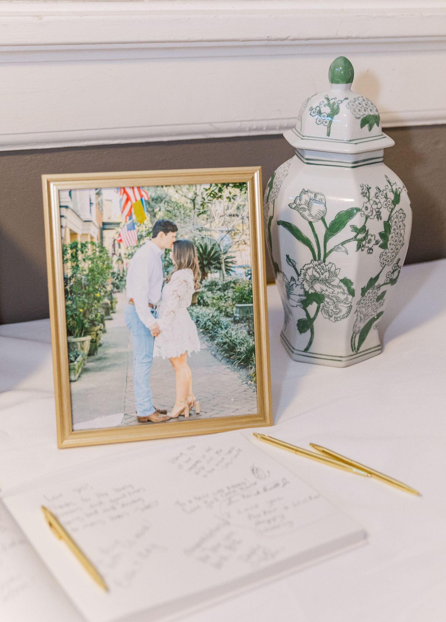 Wedding guest book with gold pen beside a framed engagement photo and decorative ginger jar vase.