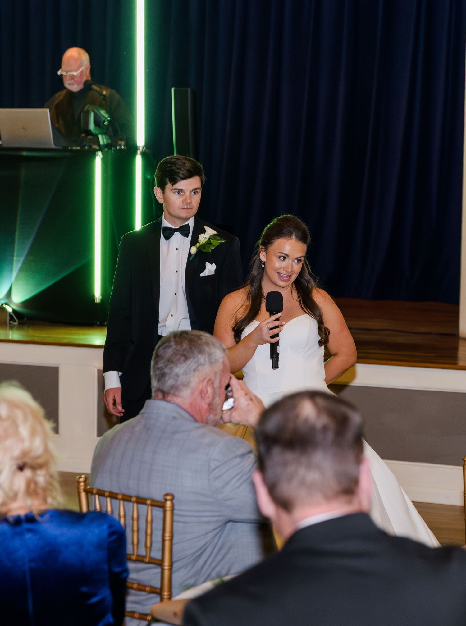 Bride speaks into microphone at wedding reception while groom stands beside her near the DJ booth.
