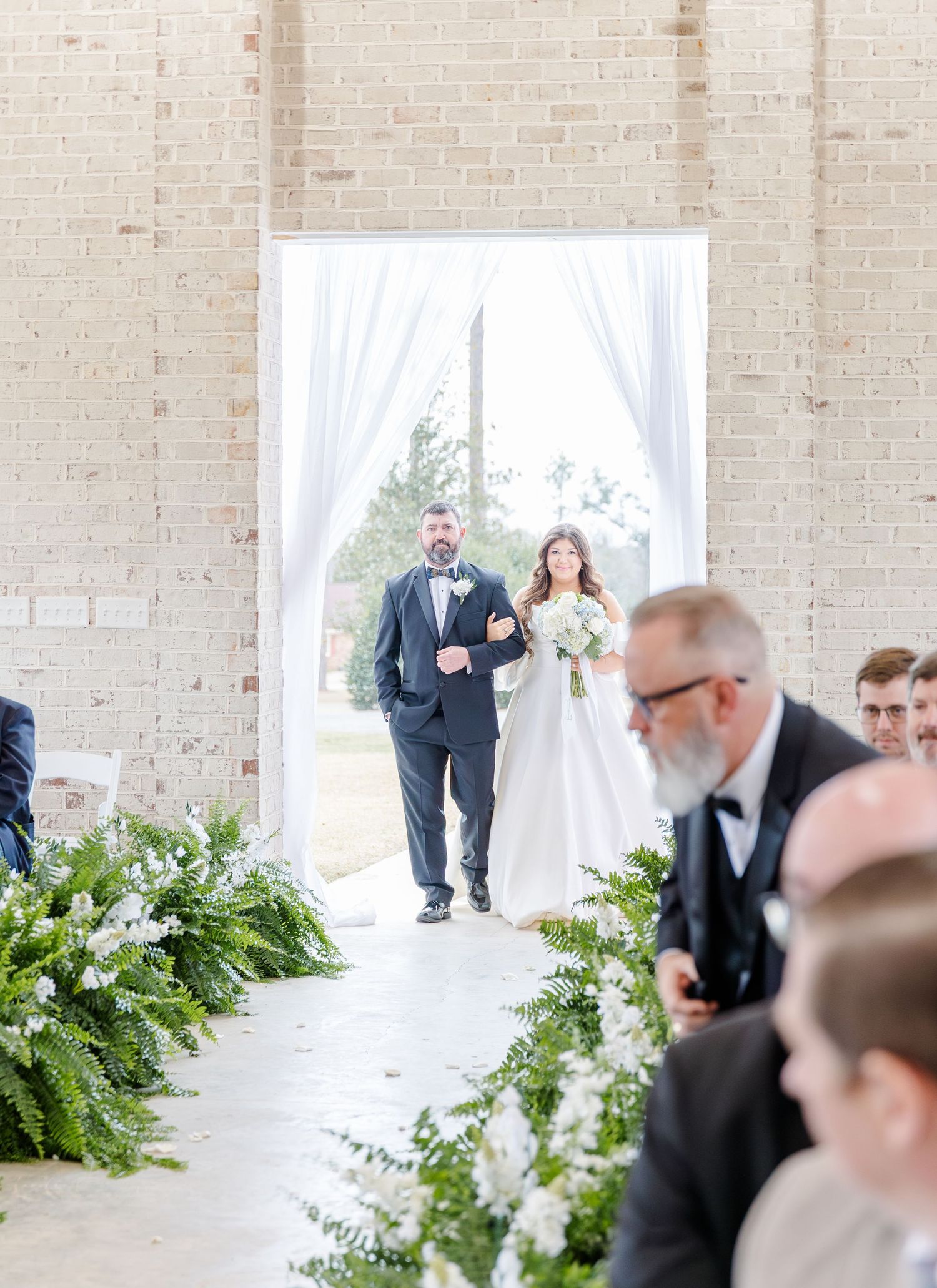 Bride walks down the aisle with her father at an elegant wedding ceremony with lush greenery and white draping.