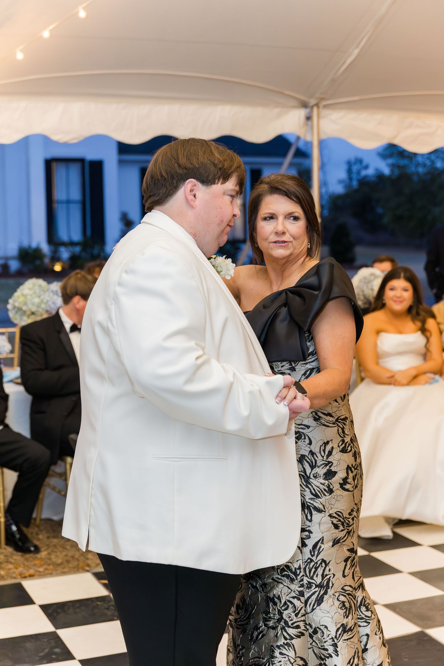 Groom in white tuxedo jacket shares mother-son dance on checkered floor under elegant wedding reception tent.