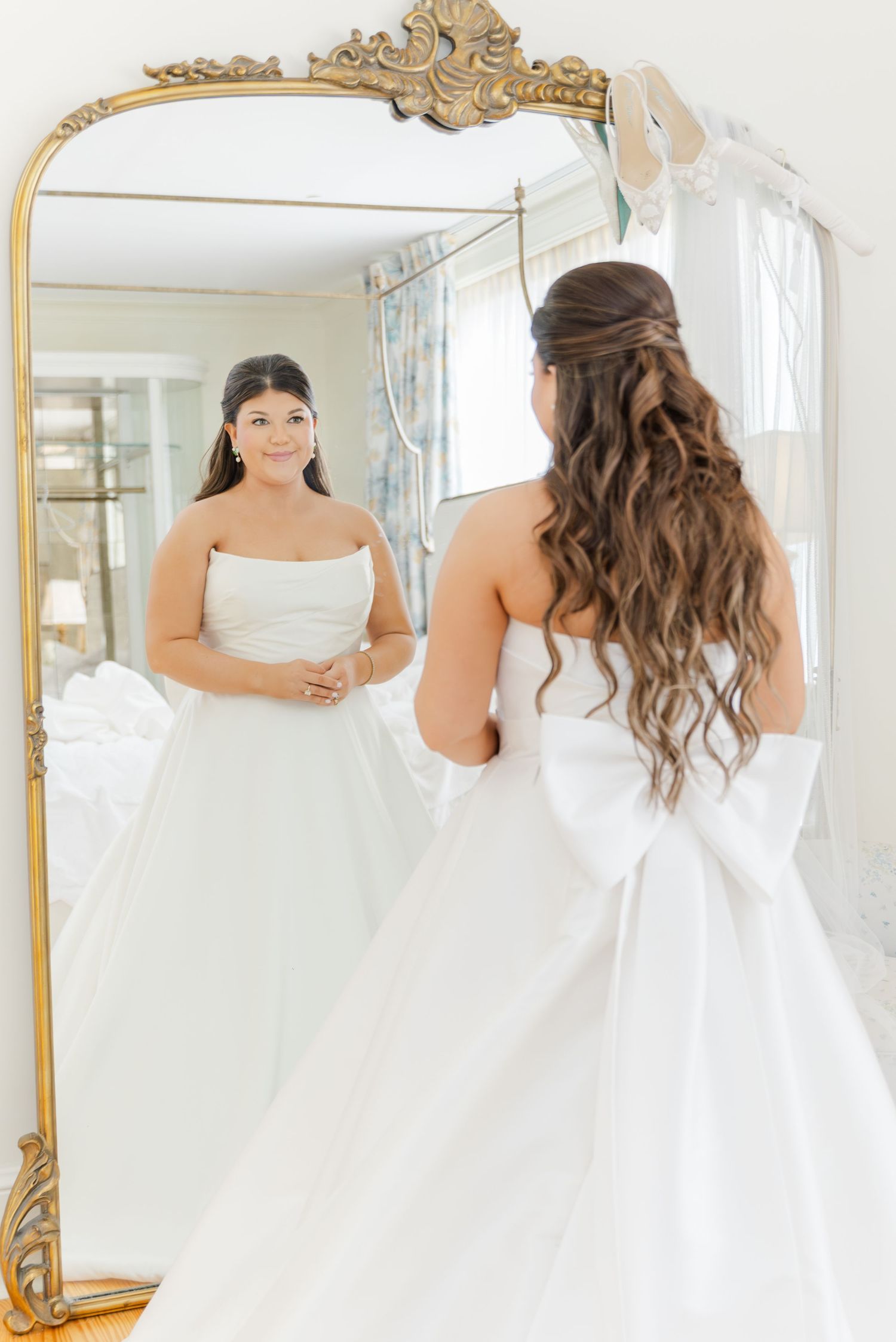 Bride in strapless white ball gown with large bow views her reflection in ornate gold mirror while getting ready.