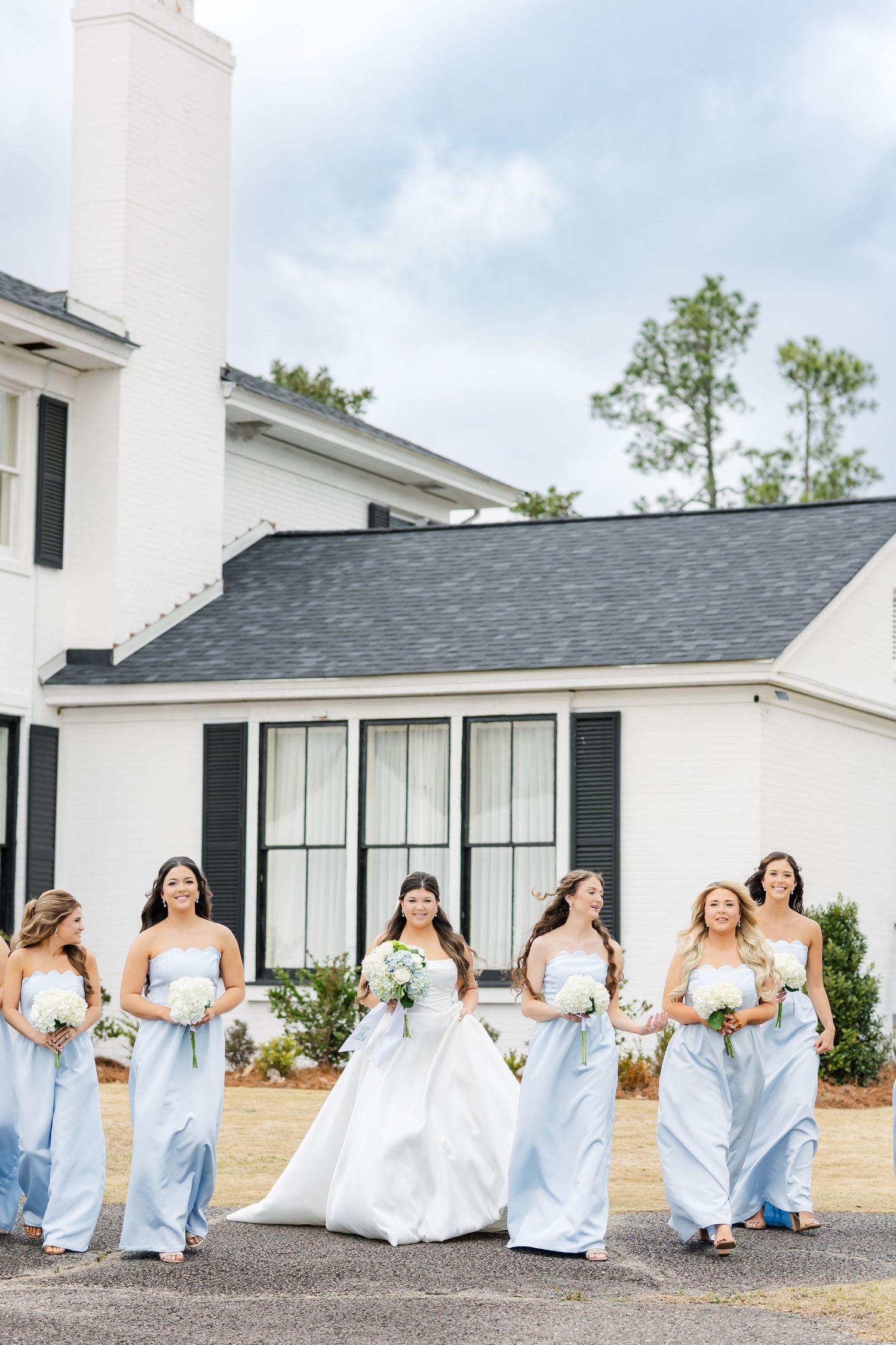 Bride in white gown walks with bridesmaids in light blue dresses outside white wedding venue.