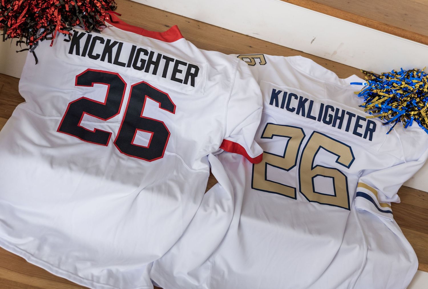Two white Kicklighter #26 football jerseys displayed side by side with blue and red pom-poms in background.