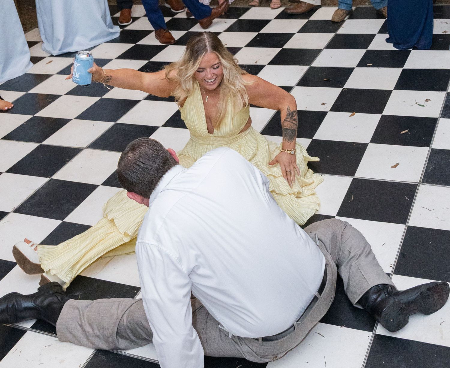 Two wedding guests playfully sitting on checkered dance floor during energetic wedding reception.