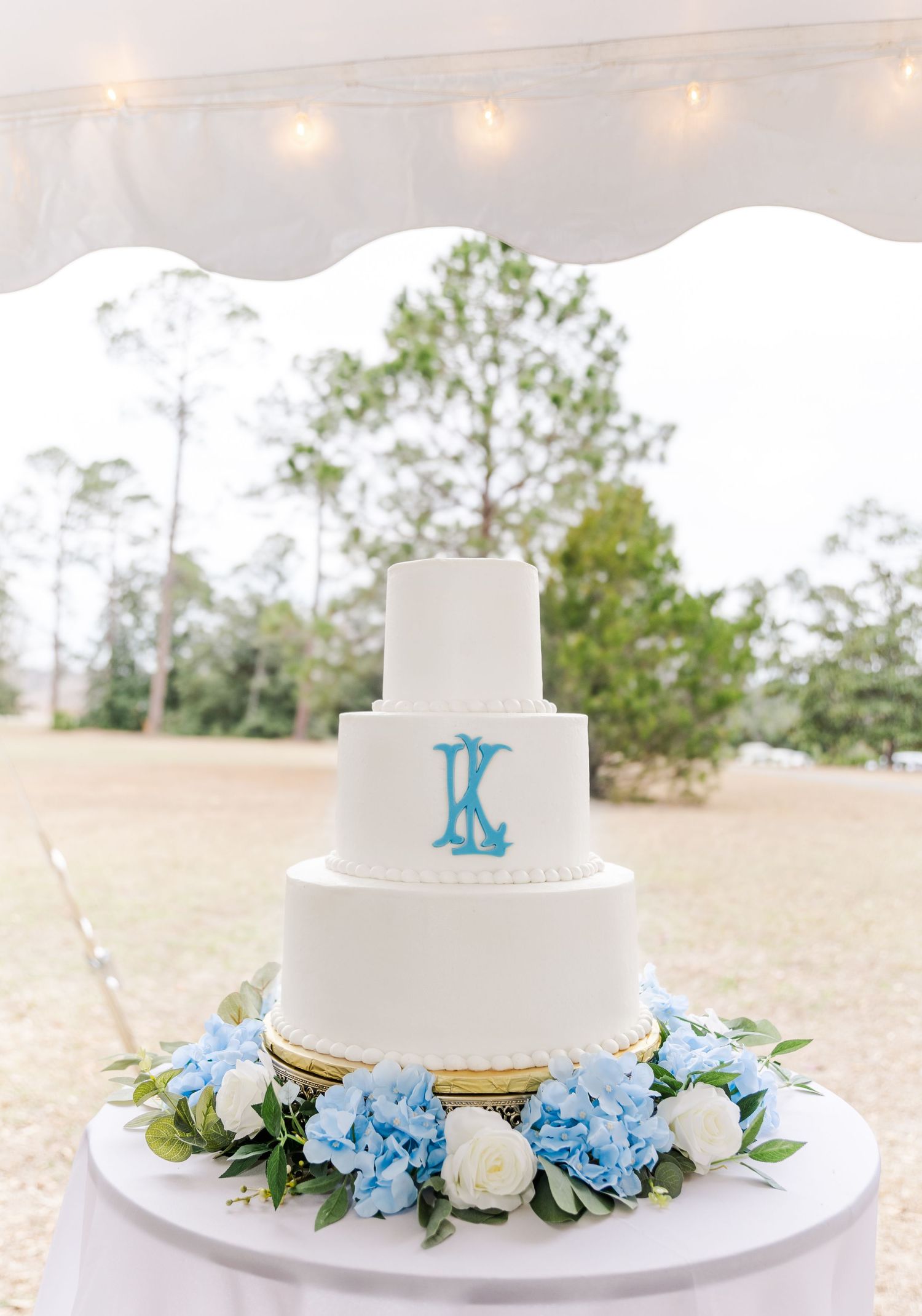 Four-tier white wedding cake with blue monogram topper surrounded by hydrangeas and white roses.
