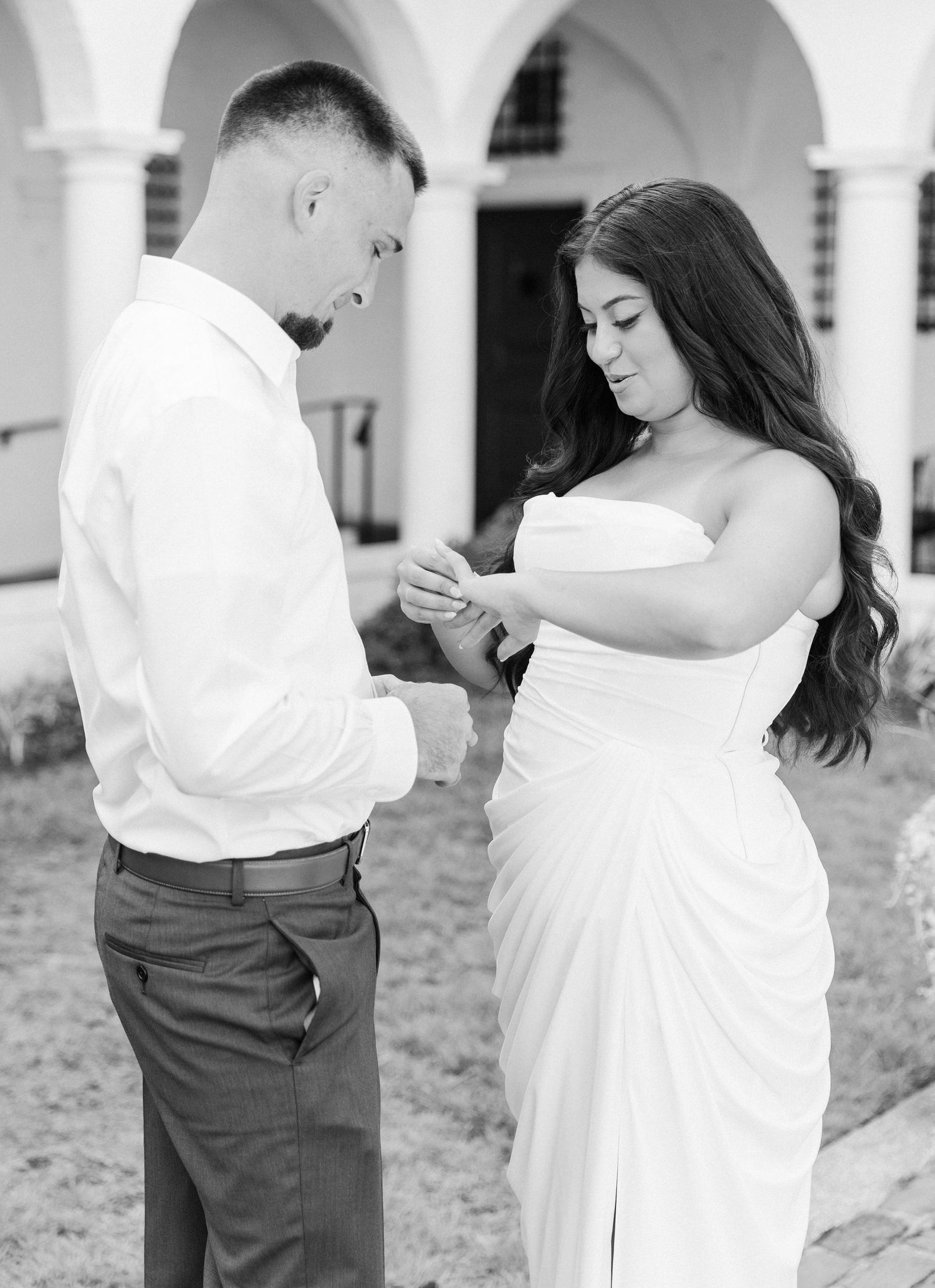 Couple examines engagement ring in black and white photo outside historic building.