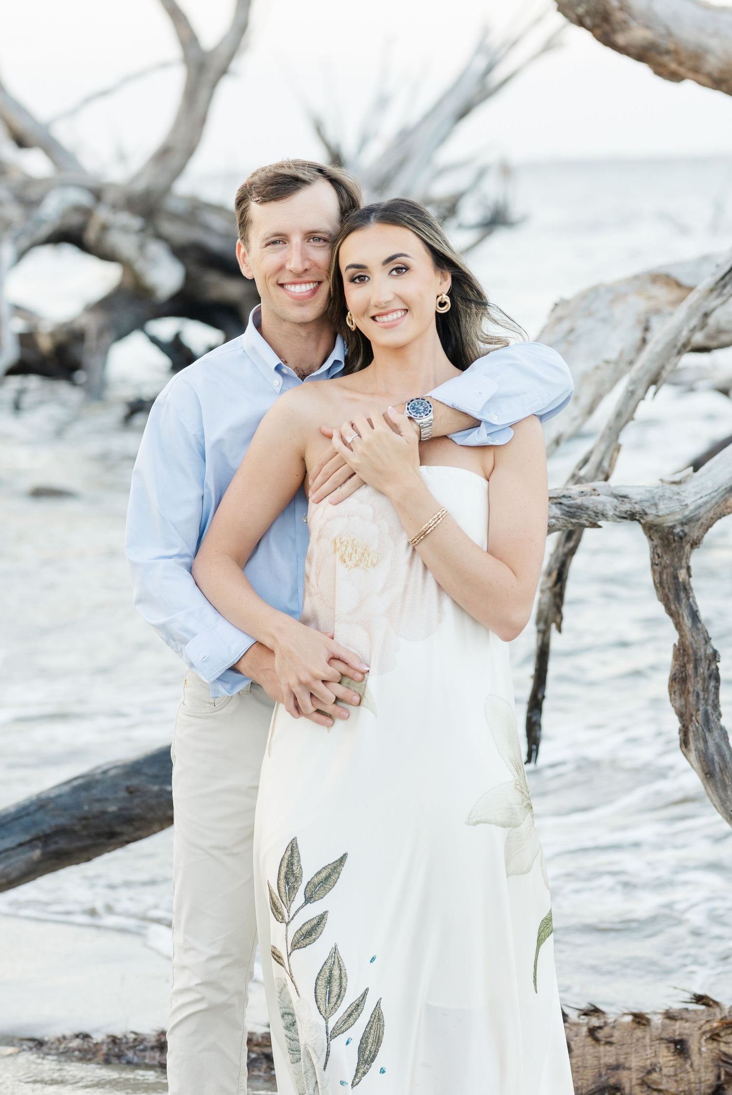 Smiling couple posing by driftwood on beach, man in blue shirt embracing woman in white floral dress.