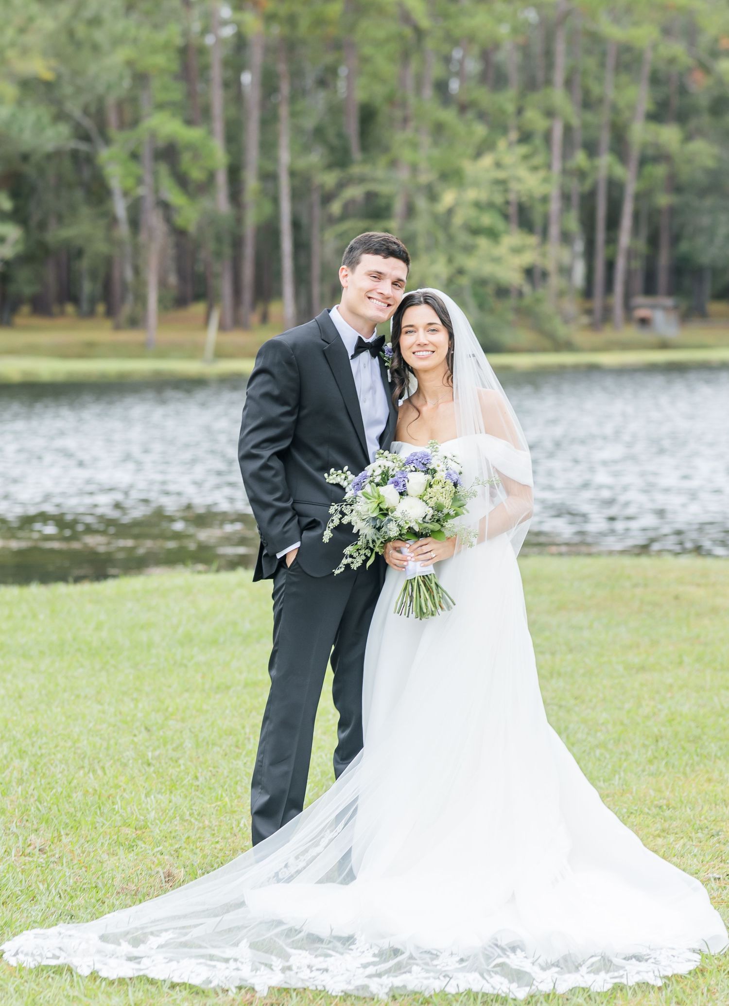 A romantic wedding couple poses together by a serene lake surrounded by lush trees and greenery.