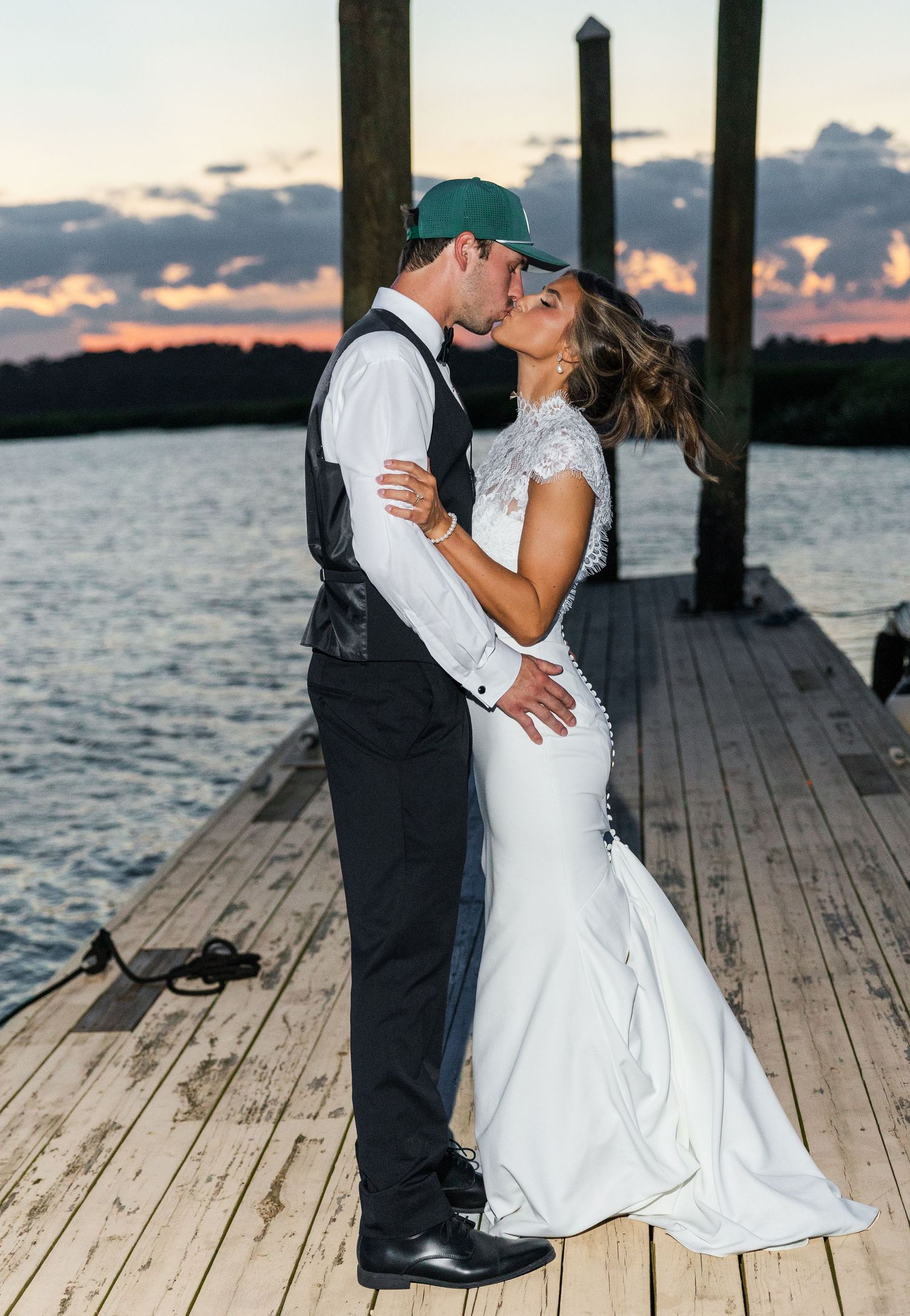Bride and groom share a romantic kiss on a waterfront dock at sunset, she in a mermaid gown, he in a vest and cap.