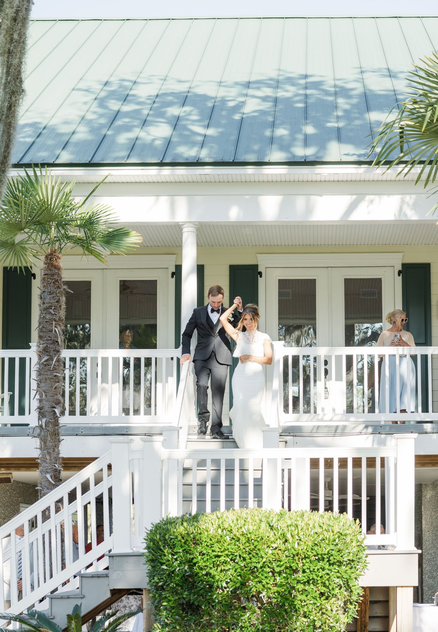 Bride and groom celebrate on a white balcony of a charming Southern venue with green shutters and palm trees.