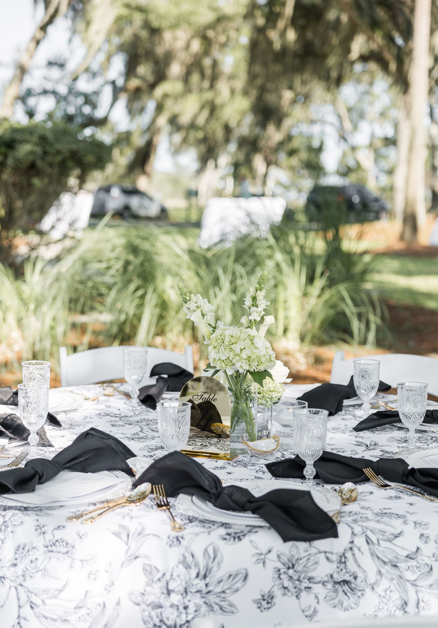 Elegant outdoor wedding table with black napkins, crystal glasses, gold cutlery, and white floral centerpiece on floral tablecloth.