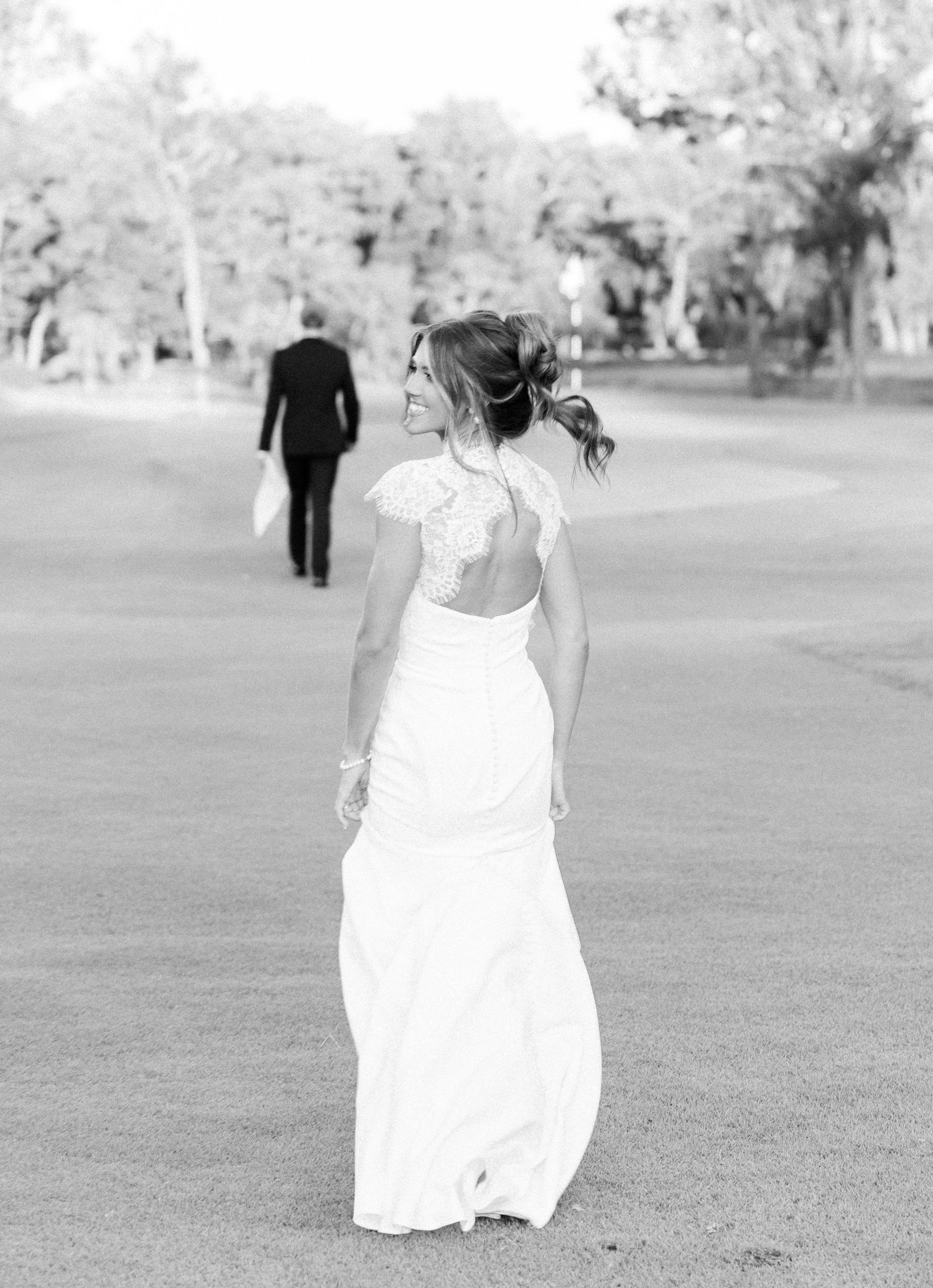 Black and white photo of a bride in a lace-back gown smiling over her shoulder as groom walks ahead.