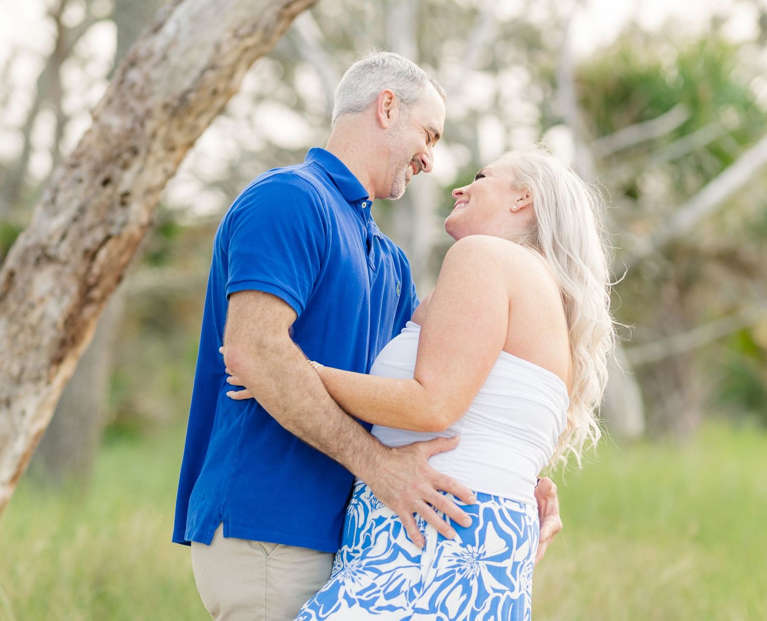 Romantic outdoor photoshoot as couple in blue and white outfits embrace and kiss against blurred nature background.