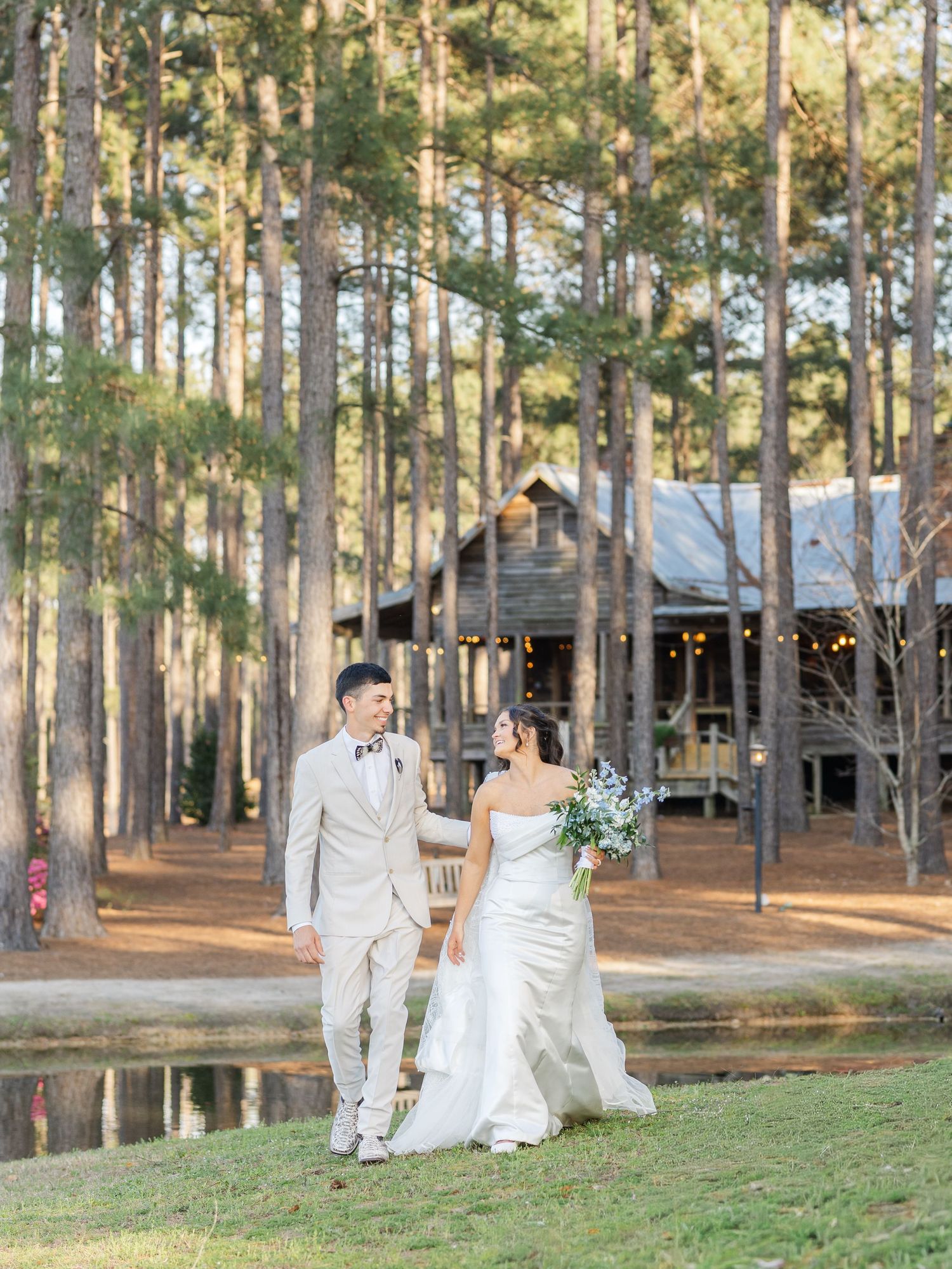 A couple in wedding attire walks together on a grassy lawn surrounded by tall pine trees and a rustic cabin.