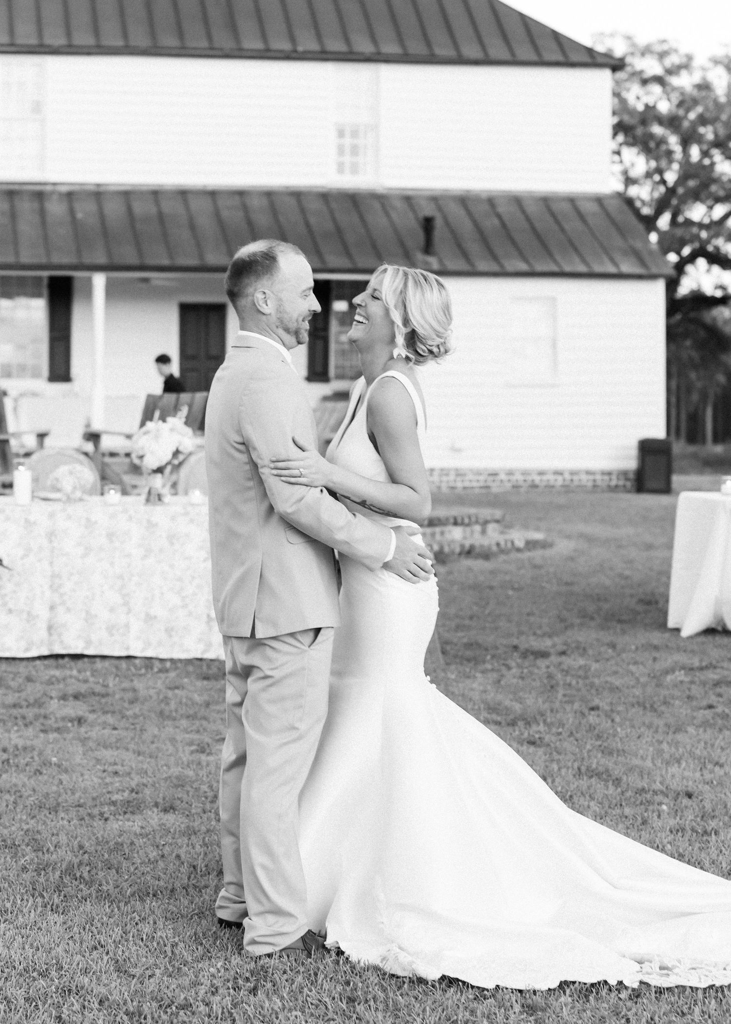 Black and white photo of laughing bride and groom embracing during outdoor wedding reception.