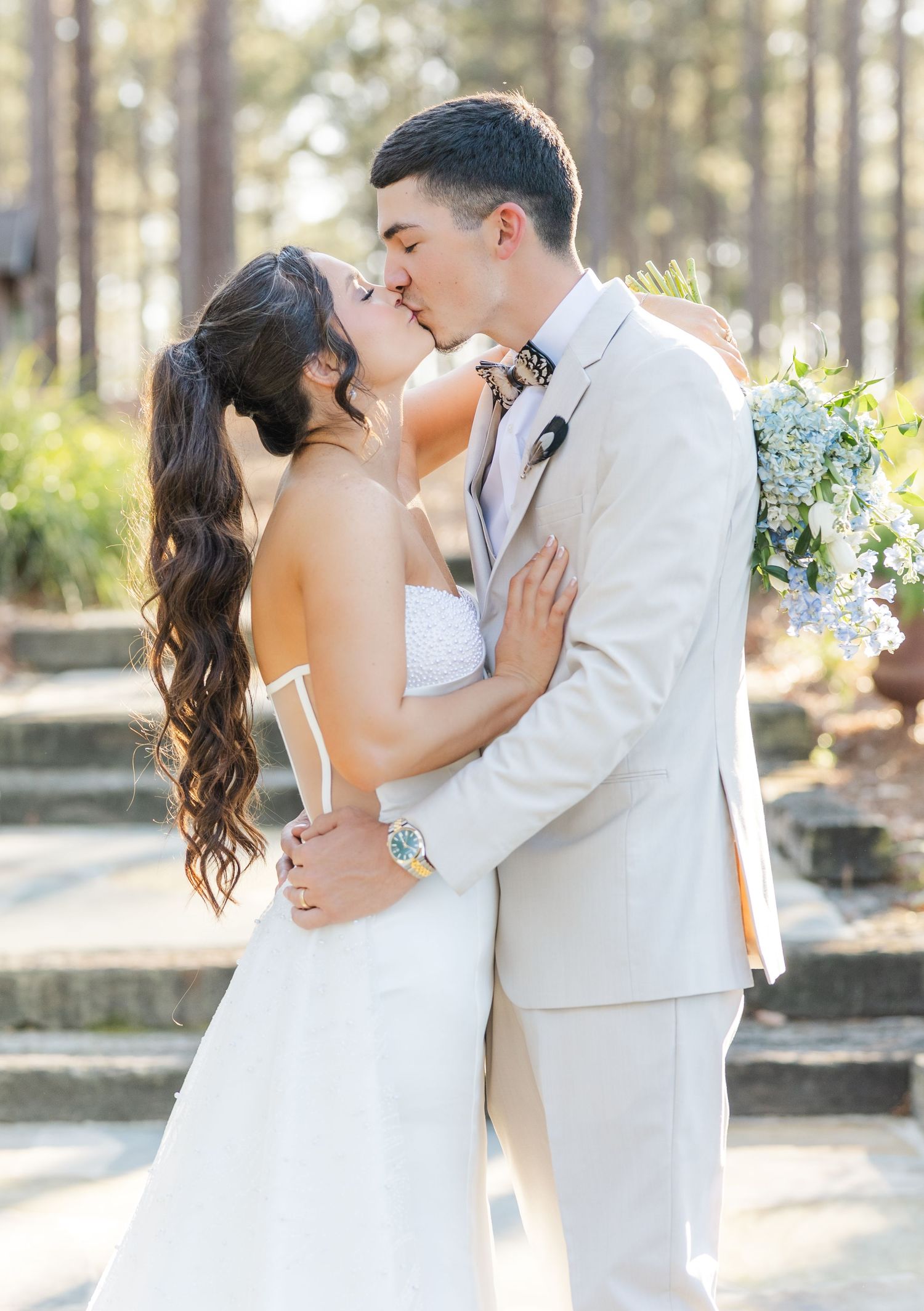 Bride and groom sharing first kiss outdoors, groom in beige suit with floral bow tie.