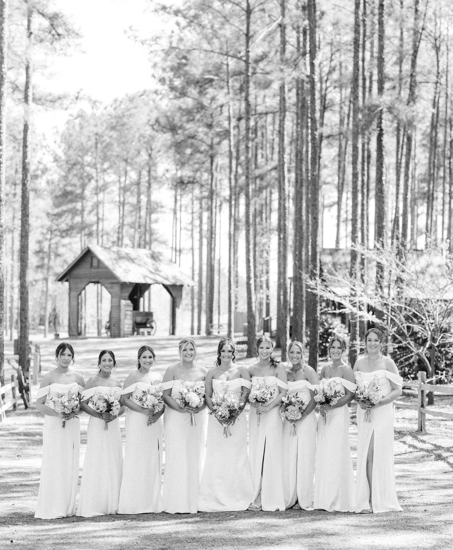 Black and white photo of bridesmaids in matching gowns holding bouquets among tall pine trees.