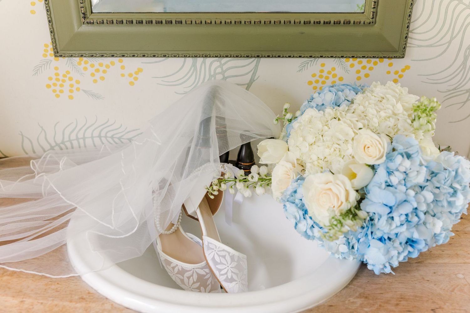 Bridal accessories flat lay with lace heels, white veil, and blue hydrangea bouquet on a marble surface.