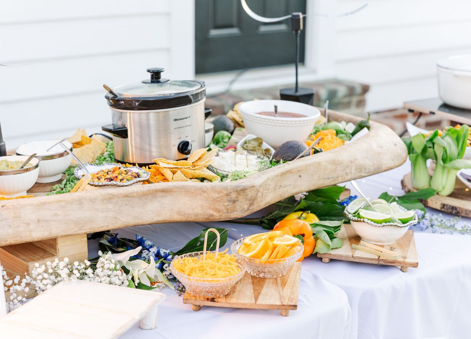 Rustic wedding food spread on white tablecloth with wooden dough bowl, slow cooker, chips, cheese, and fresh vegetables.