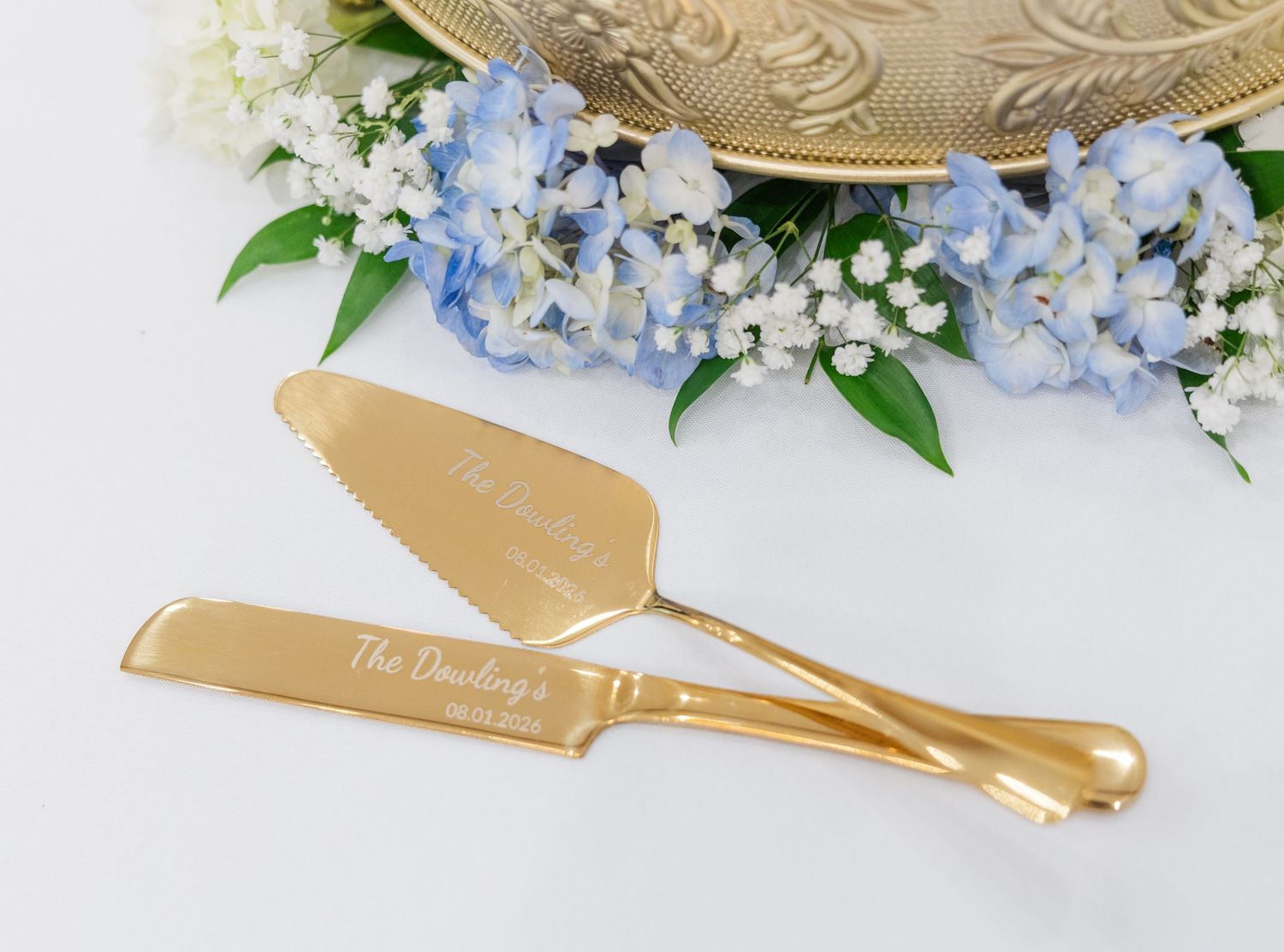 Gold personalized engraved wedding cake server set with hydrangea floral arrangement on white background.