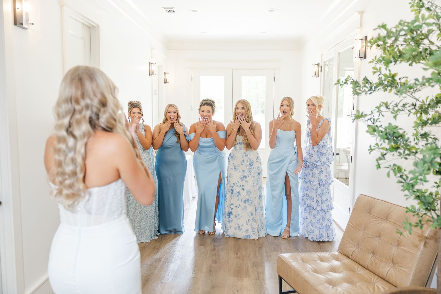 Bridesmaids in blue dresses react with excitement seeing bride in white gown for first time.