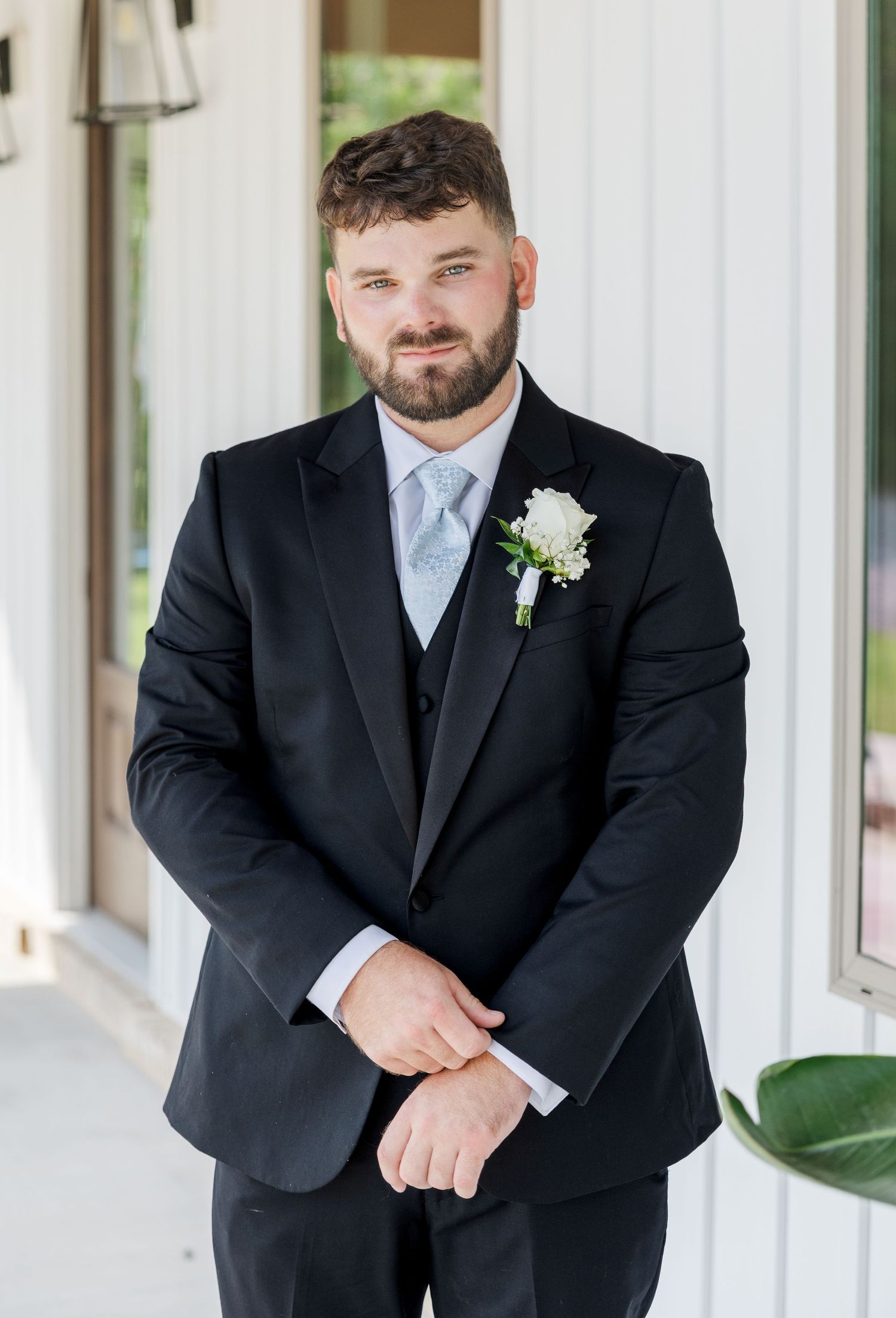 Dapper groom in black suit with white floral boutonniere and blue tie smiling confidently at wedding venue.