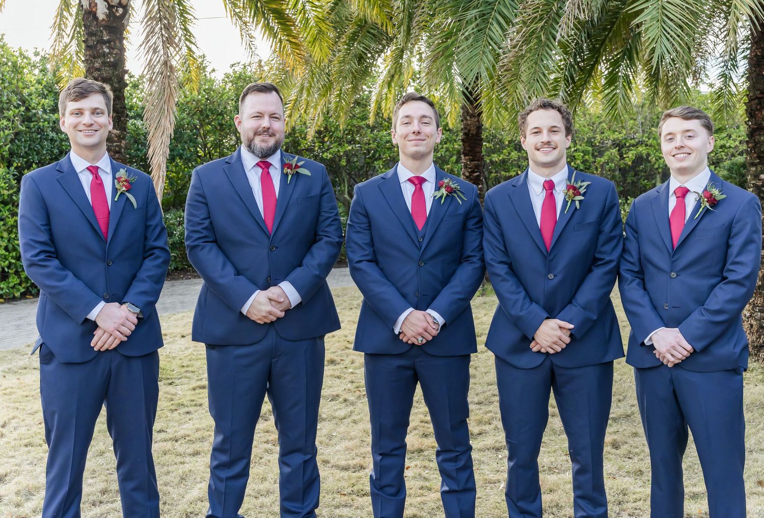 Groomsmen in navy suits with red ties standing in a row outdoors.