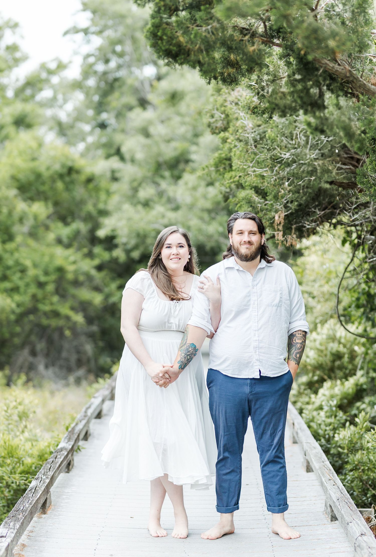 Romantic couple sitting barefoot on wooden boardwalk surrounded by lush green foliage.