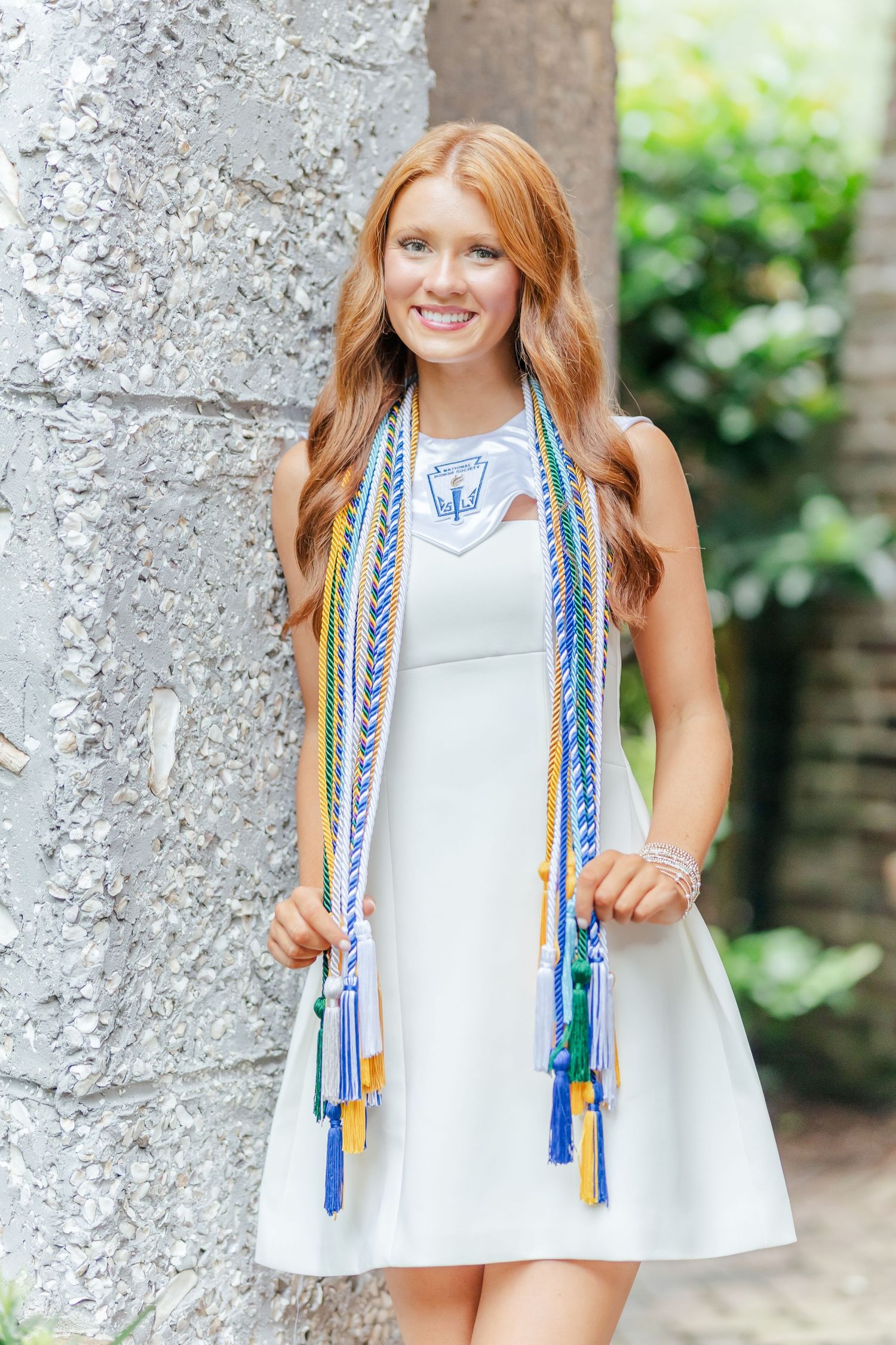 Woman wearing white dress with decorative blue and white tasseled trim stands against stone wall.