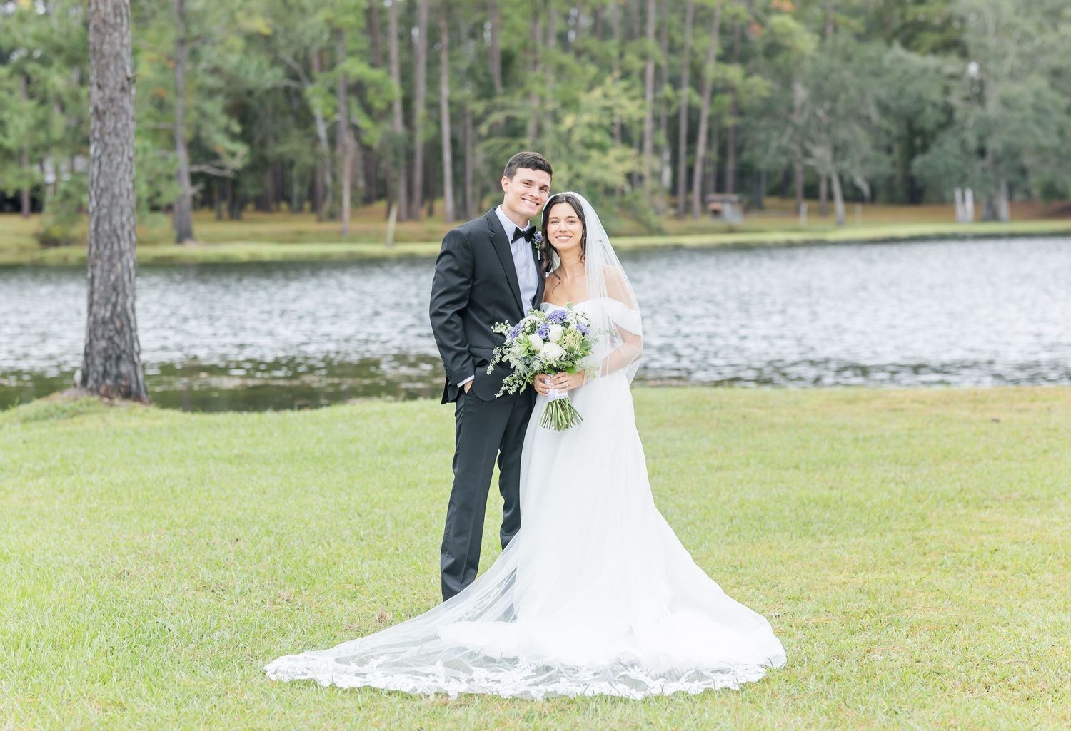Newlyweds pose for portrait on lawn with lake and pine trees in background.