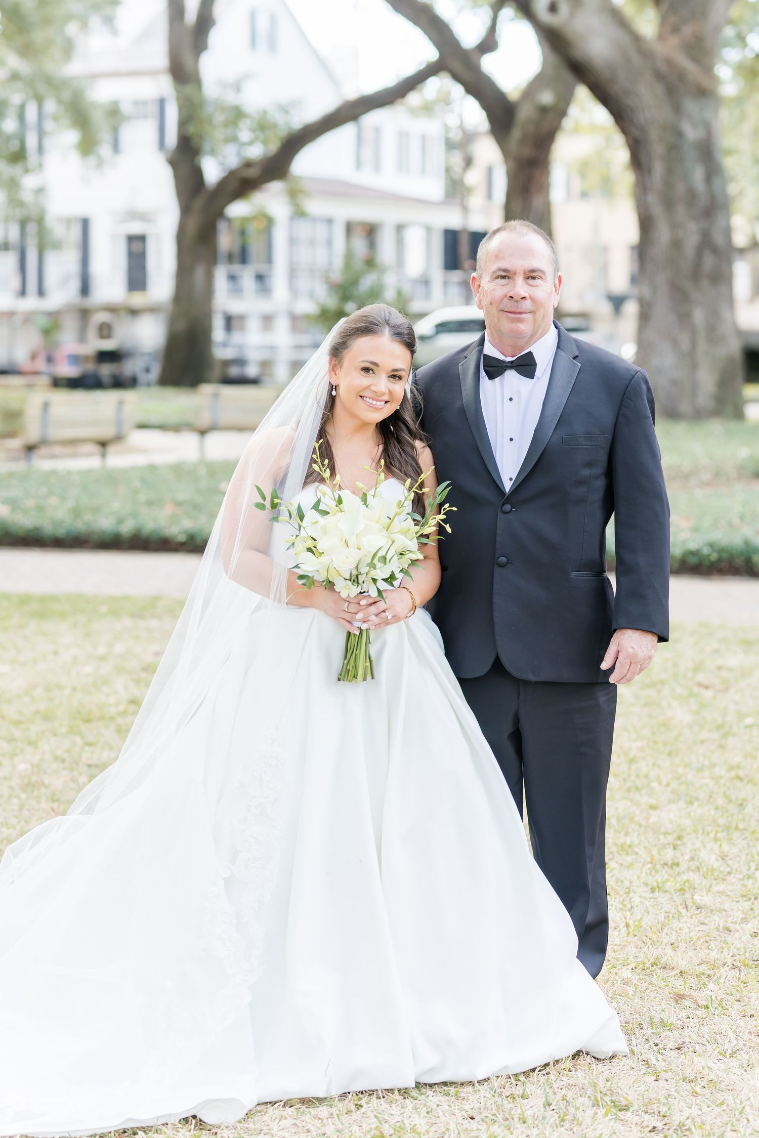 Bride in ball gown with veil holds white bouquet beside man in black tuxedo at outdoor wedding.