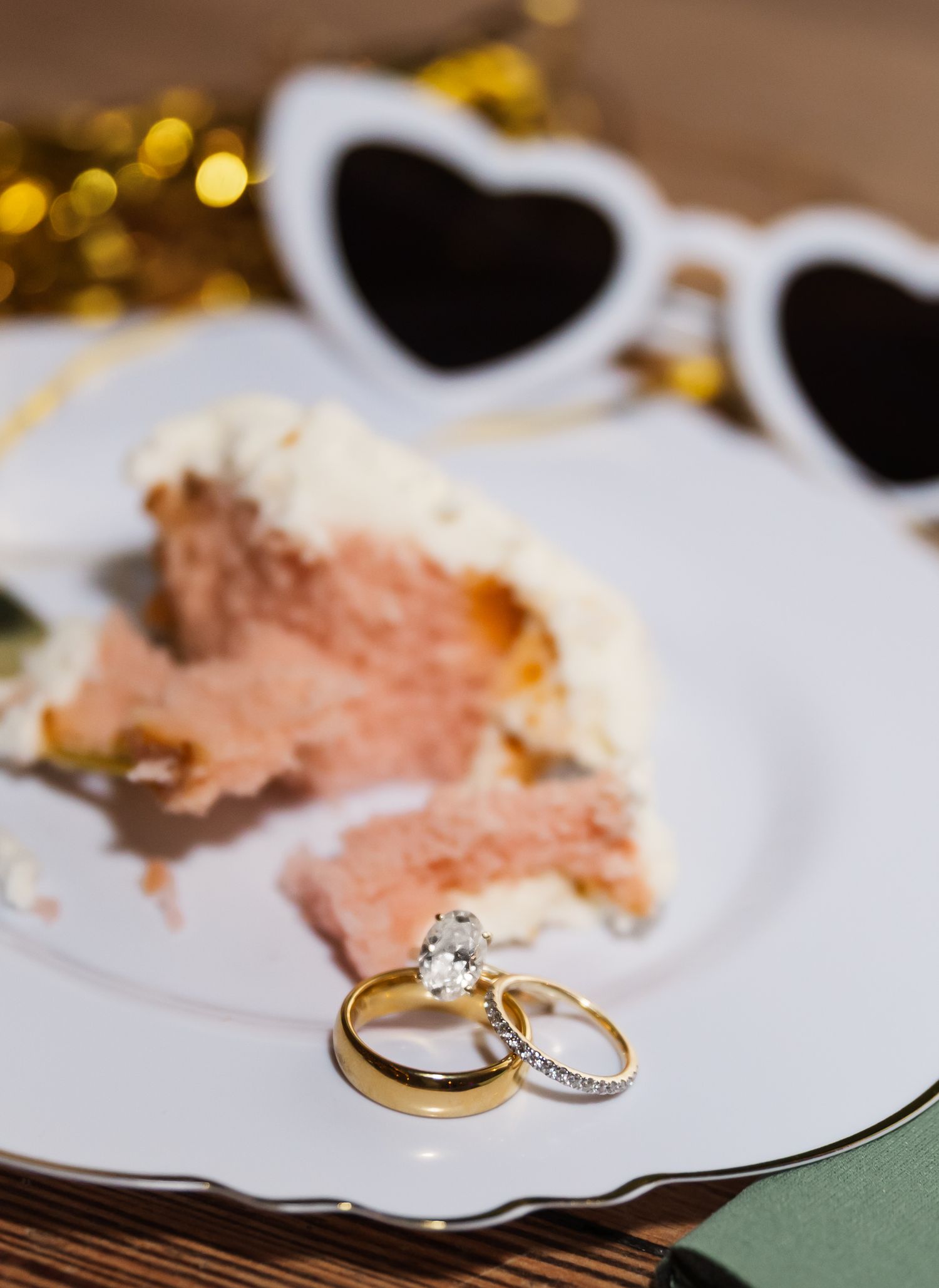 Wedding rings and diamond engagement ring displayed on white plate with pink cake slice and heart sunglasses.