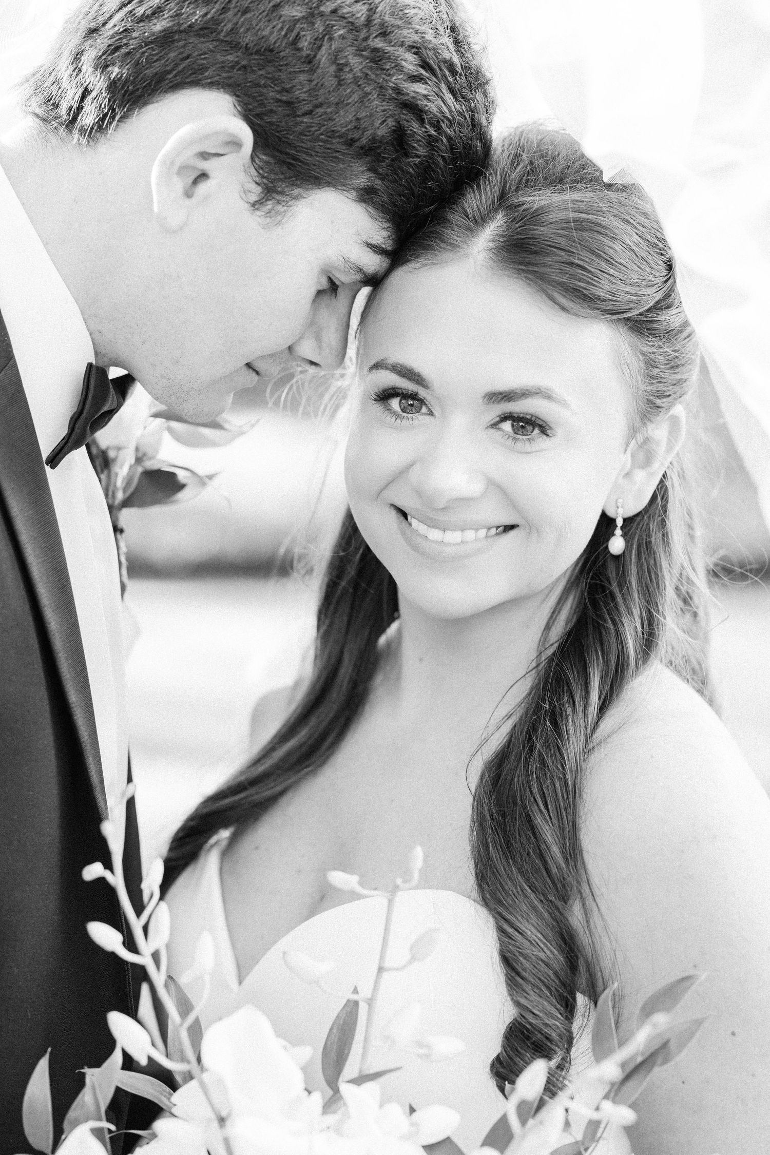 Black and white close-up of smiling bride leaning against groom in tuxedo holding white floral bouquet.
