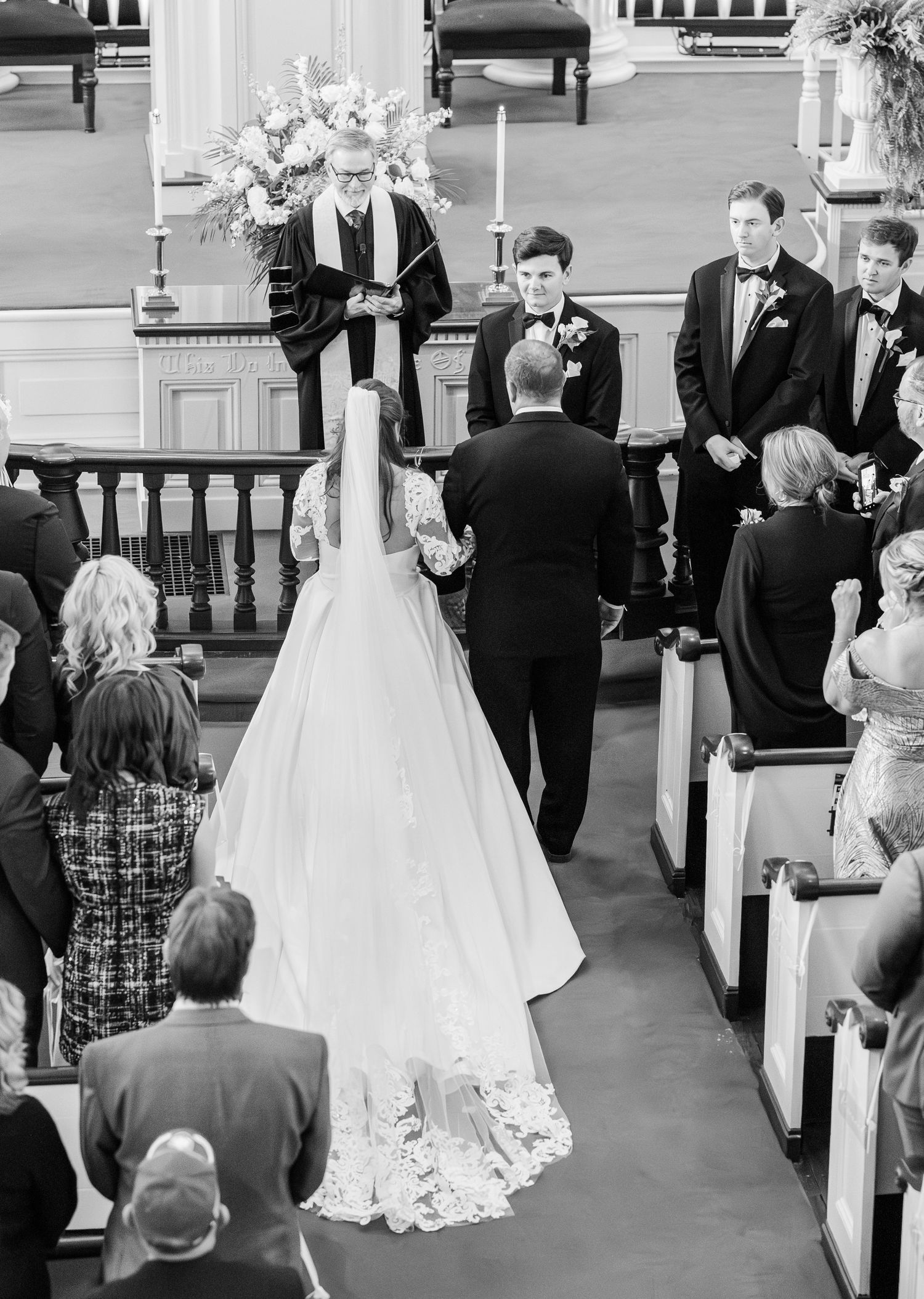 Black and white wedding ceremony photo of bride and groom at church altar with officiant and groomsmen.