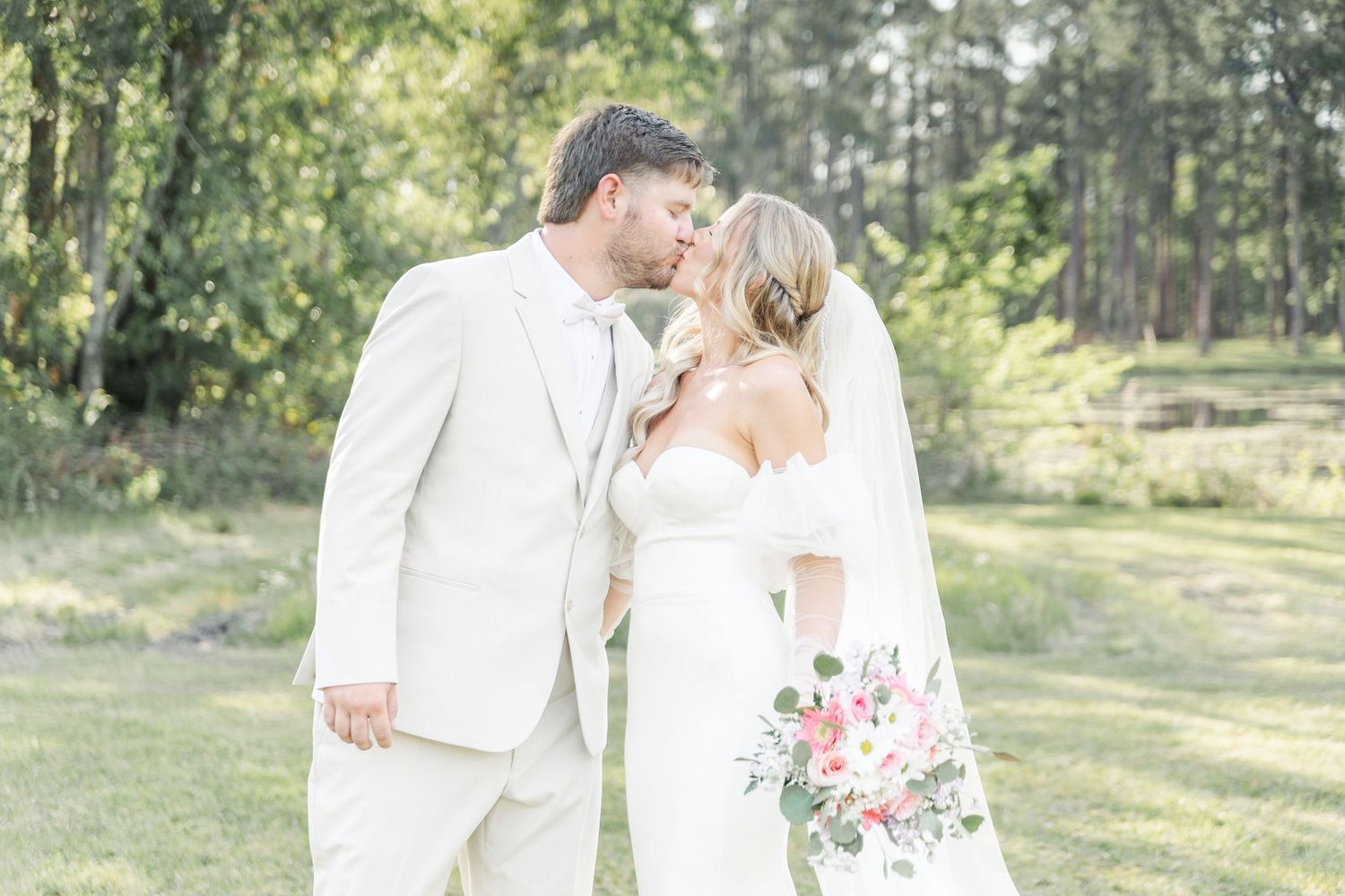 Wedding couple in white attire share an intimate moment on their wedding day with forest scenery behind them.