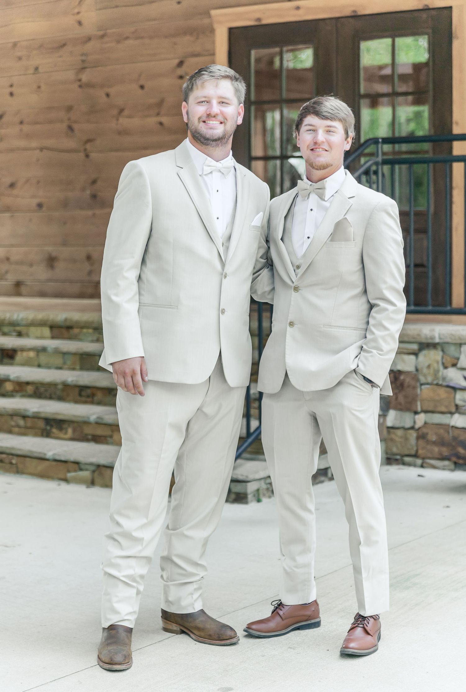 Two figures in matching light suits and bow ties stand on a concrete patio against wooden cabin siding.
