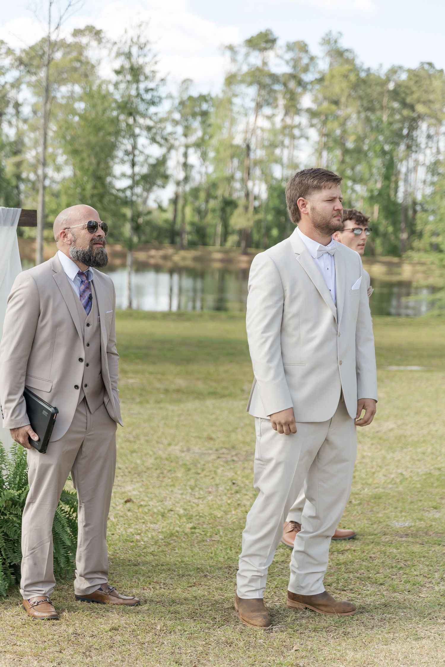 Groomsmen in light colored suits stand on grass with a pond in the background.