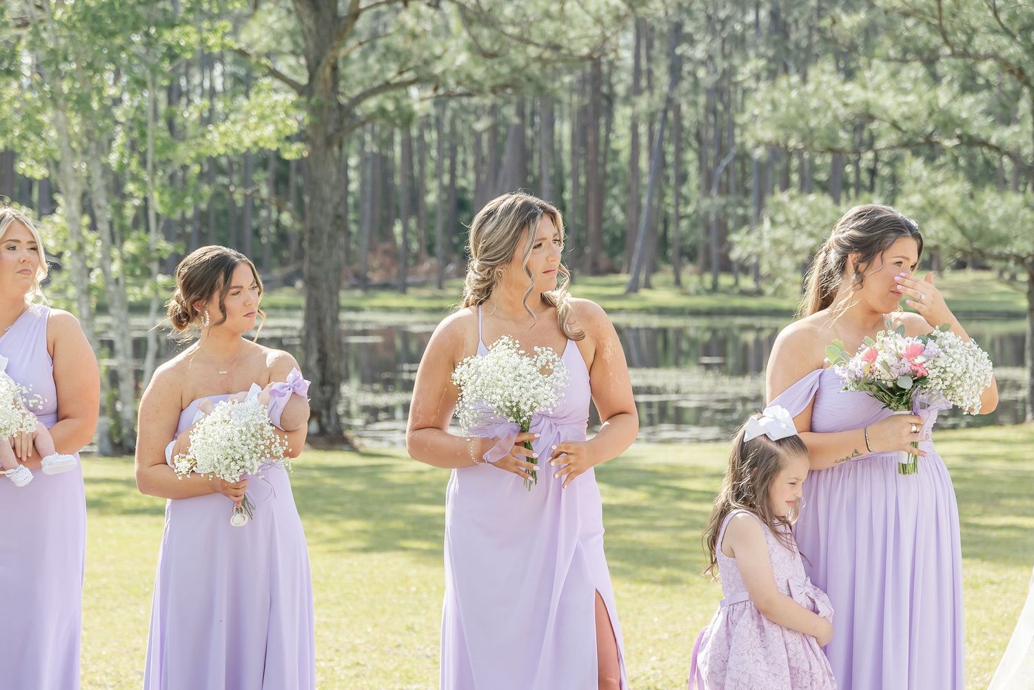 Bridesmaids wearing flowing lavender dresses hold bouquets during outdoor wedding ceremony by a lake.