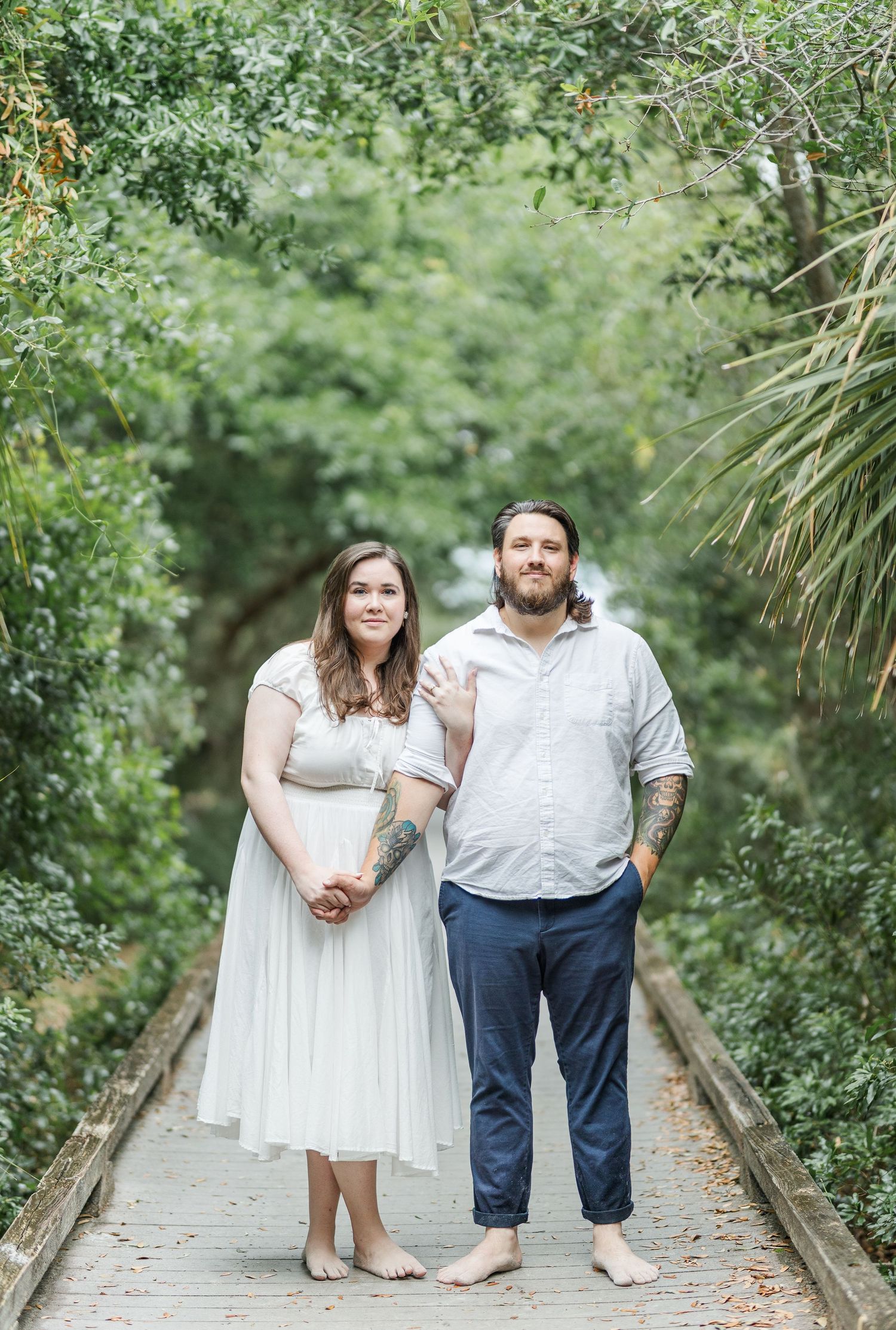 A couple stands barefoot together on a wooden boardwalk surrounded by lush greenery.