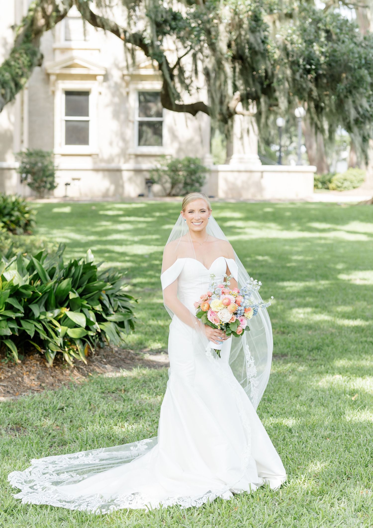 A bride in a white strapless wedding gown poses gracefully on a manicured lawn with Spanish moss trees in the background.