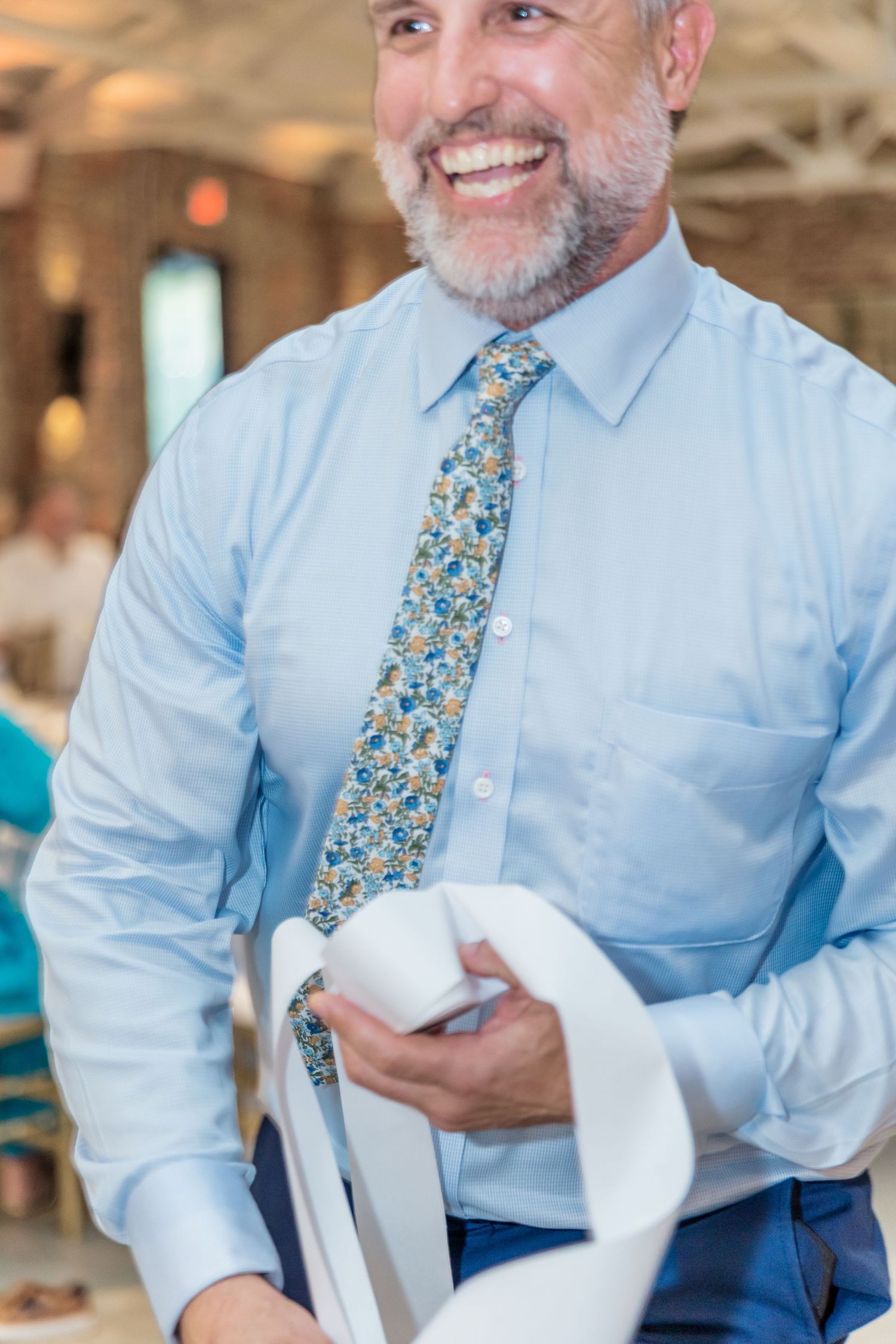 Person in light blue dress shirt and floral tie smiles during wedding reception.