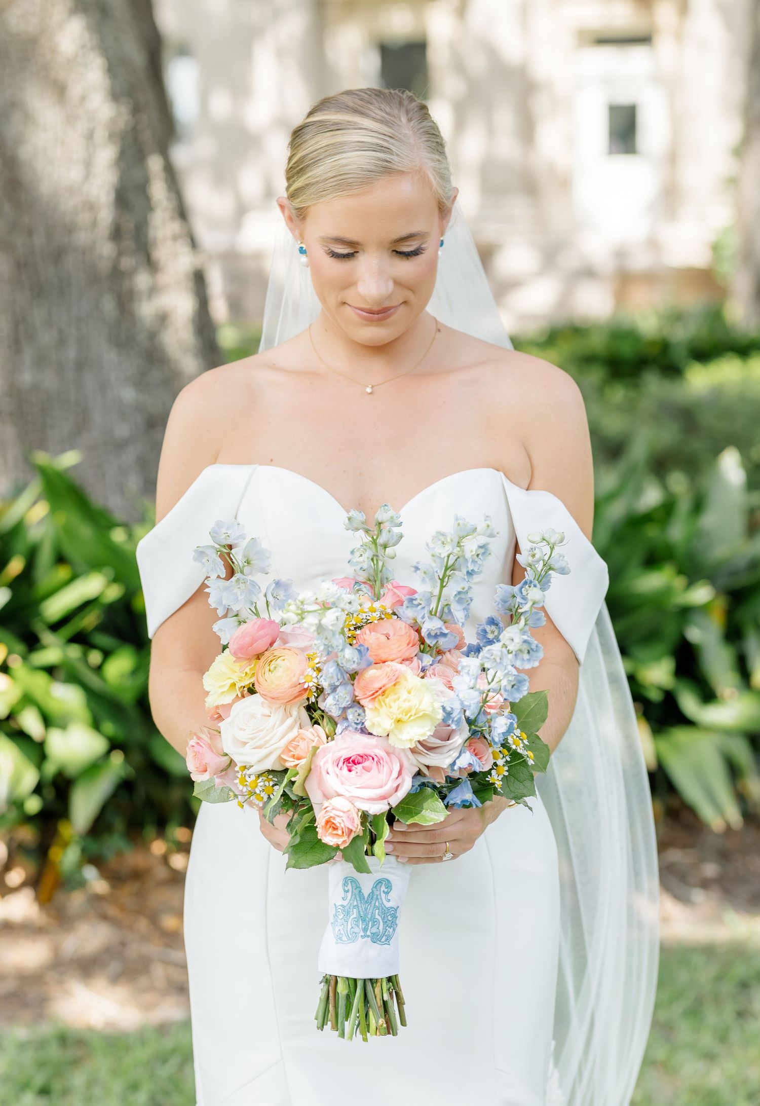 A bride in a white off-shoulder gown holds a vibrant bouquet of peach, pink and blue flowers in a garden setting.